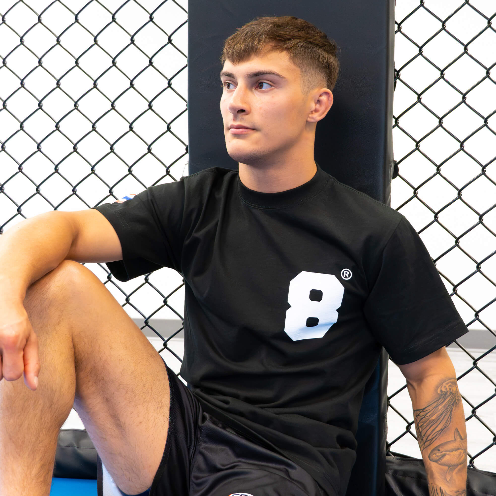 A young male fighter with an arm tattoo sits relaxed against an MMA cage corner pad, wearing the 8 Thai Boxing T-Shirt with a white block number "8" on the chest.