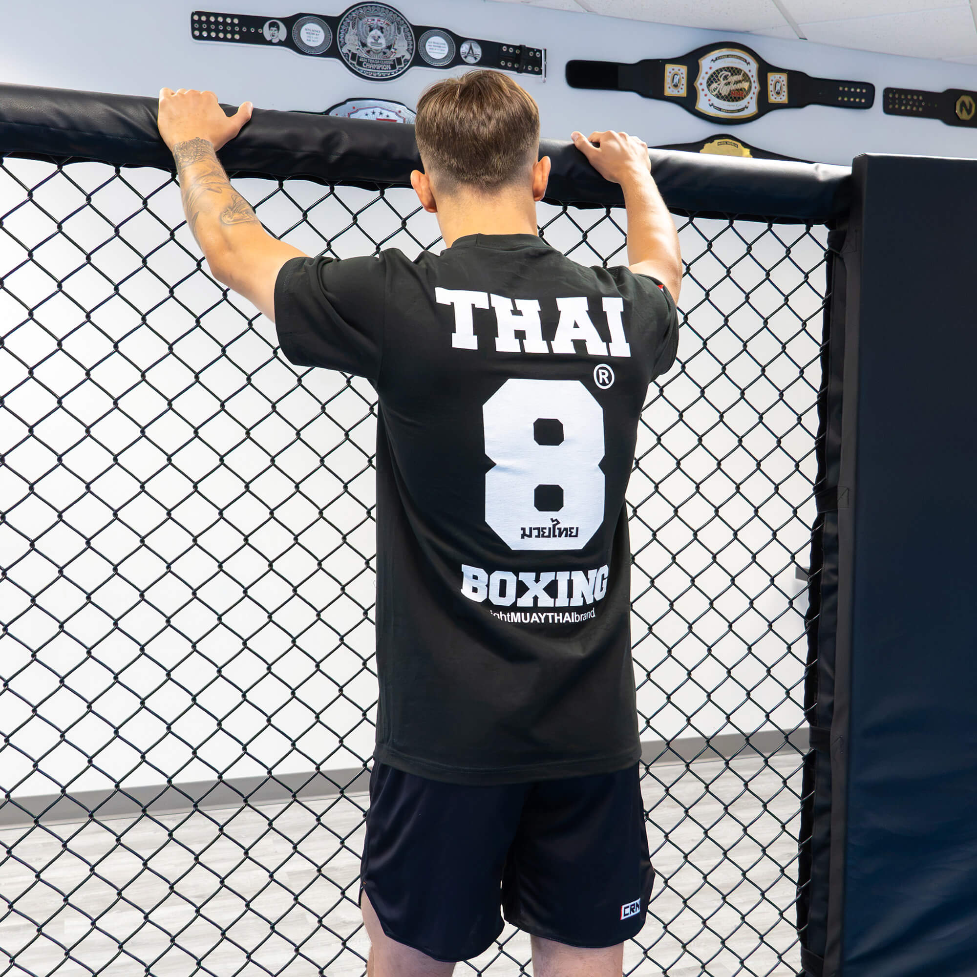 Rear view of a male athlete standing at an MMA cage fence, wearing a the 8 Thai Boxing T-Shirt with large white text and a bold number eight graphic on the back.