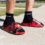 A close-up lifestyle shot of a person's feet wearing Combat Corner CRNR Foam Slides in the red and black Cinder Topo camouflage pattern. The slides are paired with black mid-calf socks that feature the white "CRNR" logo stitched near the ankle cuff. The model is standing outdoors on a textured concrete pavement, showcasing the slides from a low three-quarter angle that highlights the thick foam upper, the small side "CRNR" logo badge, and the comfortable fit.