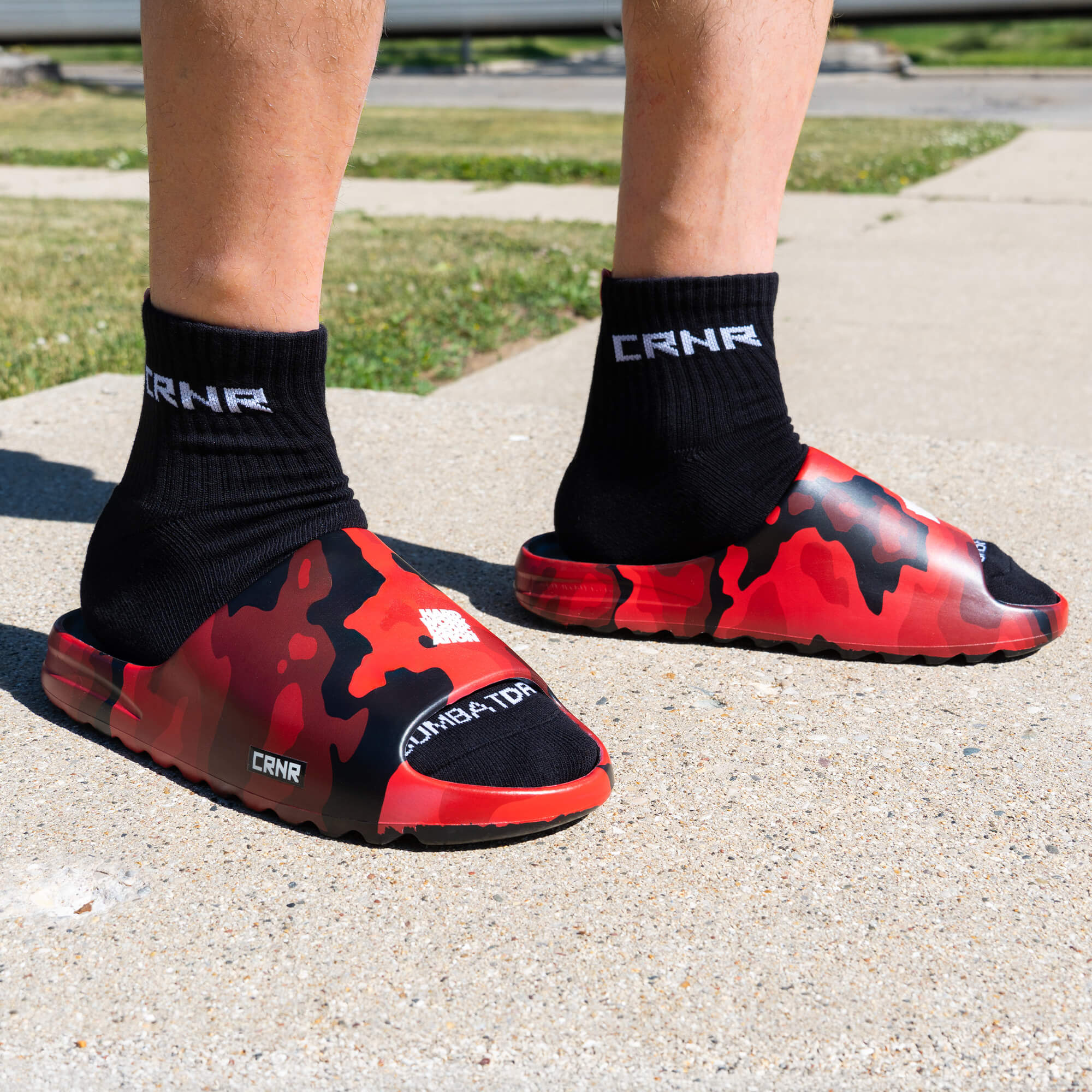 A close-up lifestyle shot of a person's feet wearing Combat Corner CRNR Foam Slides in the red and black Cinder Topo camouflage pattern. The slides are paired with black mid-calf socks that feature the white "CRNR" logo stitched near the ankle cuff. The model is standing outdoors on a textured concrete pavement, showcasing the slides from a low three-quarter angle that highlights the thick foam upper, the small side "CRNR" logo badge, and the comfortable fit.