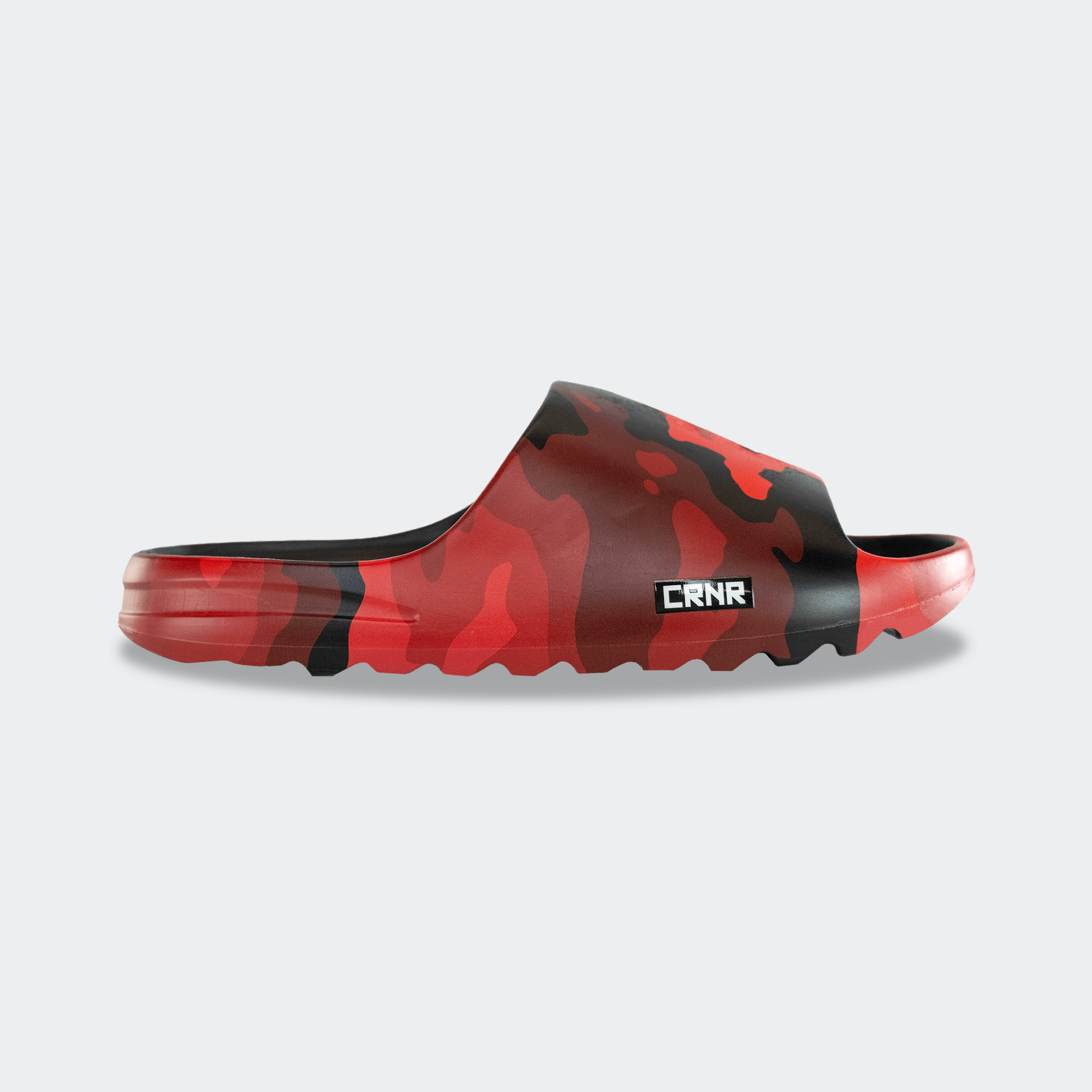 Side profile view of the outer side of a Combat Corner CRNR Foam Slide. The slide features a red and black Cinder Topo camo pattern with a small rectangular black badge displaying the white "CRNR" logo on the lower side of the strap. The sole has a thick, wavy, serrated tread pattern.