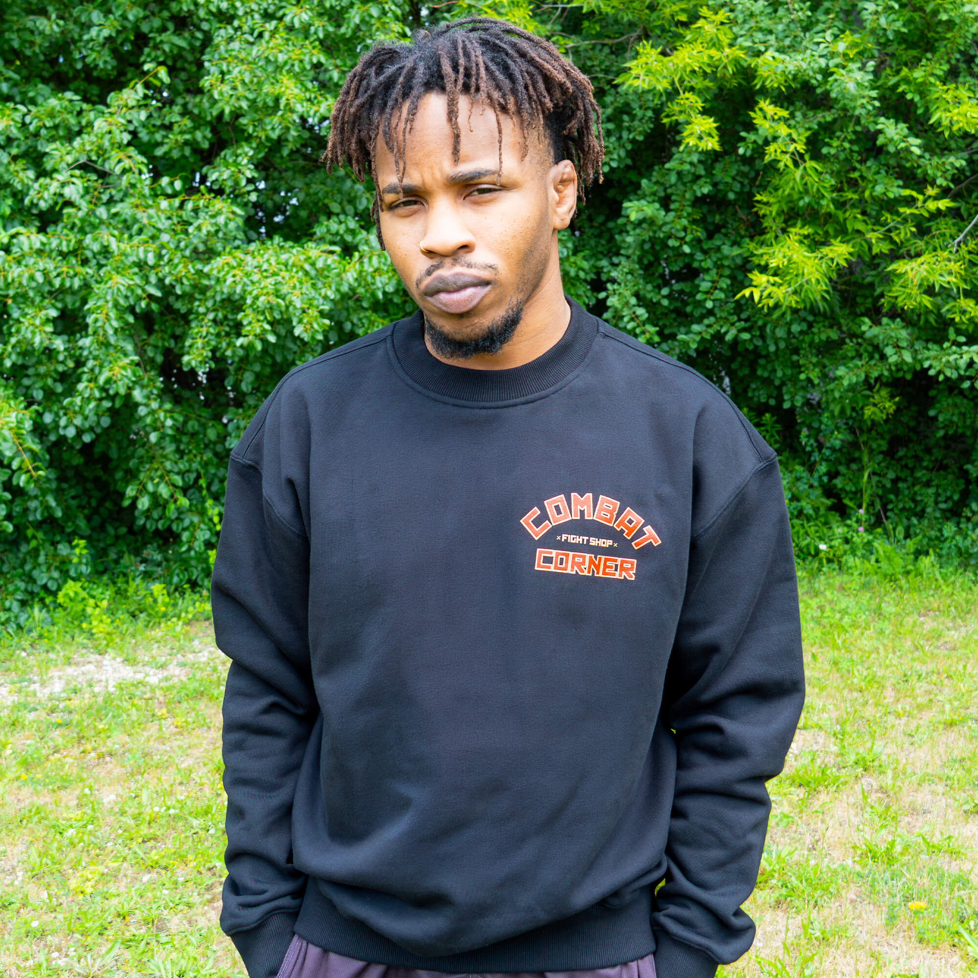 Front view of a man with dreadlocks standing outdoors in front of a green leafy background. He is wearing a black Combat Corner crewneck sweatshirt that features a small orange and red block-letter logo on the left chest reading "Combat Fight Shop Corner".
