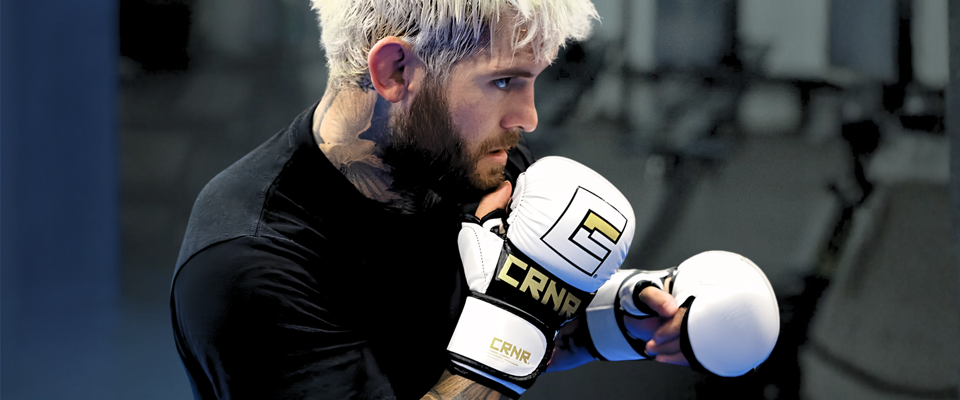 A side profile shot of a focused, tattooed male fighter with bleached hair holding up a sparring guard, wearing Combat Corner's white MMA Spar Gloves.