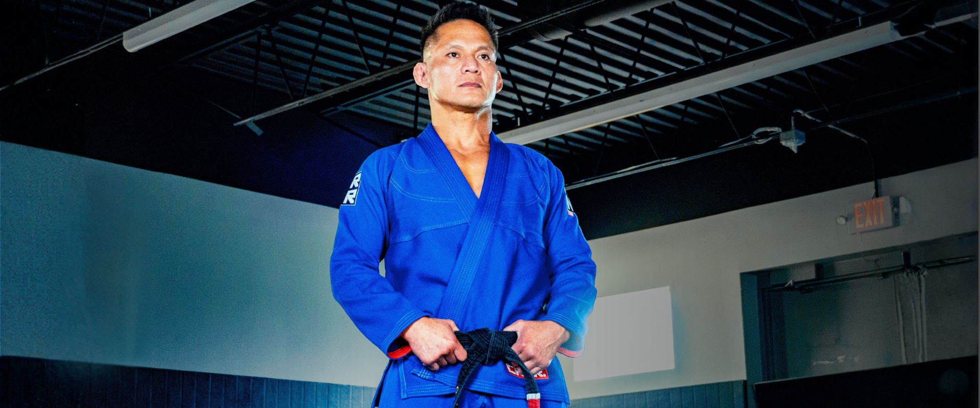 Low-angle portrait of BJJ World Champion Mark Vives standing in a gym, wearing Combat Corner’s blue V7 BJJ Gi with a black belt. 