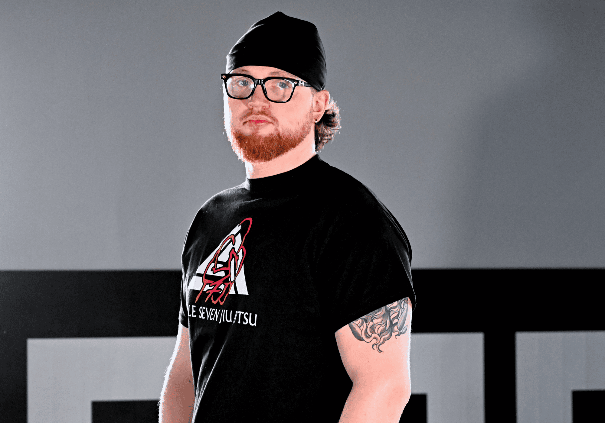 Medium shot of a man with a red beard wearing a black custom Combat Corner x Le Seven Jiu Jitsu t-shirt and a black beanie, looking toward the camera against a grey background.