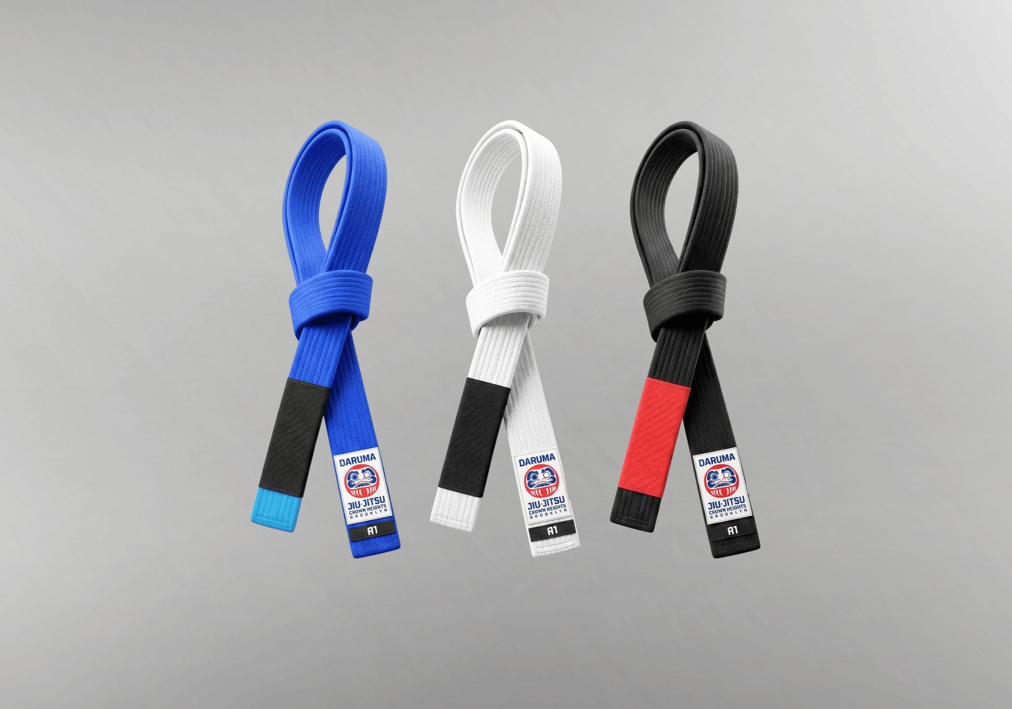 A collection of three custom Jiu Jitsu gi belts by Combat Corner in blue, white, and black. Each is knotted and features a custom "Daruma Jiu-Jitsu" patch and ranking sleeve.