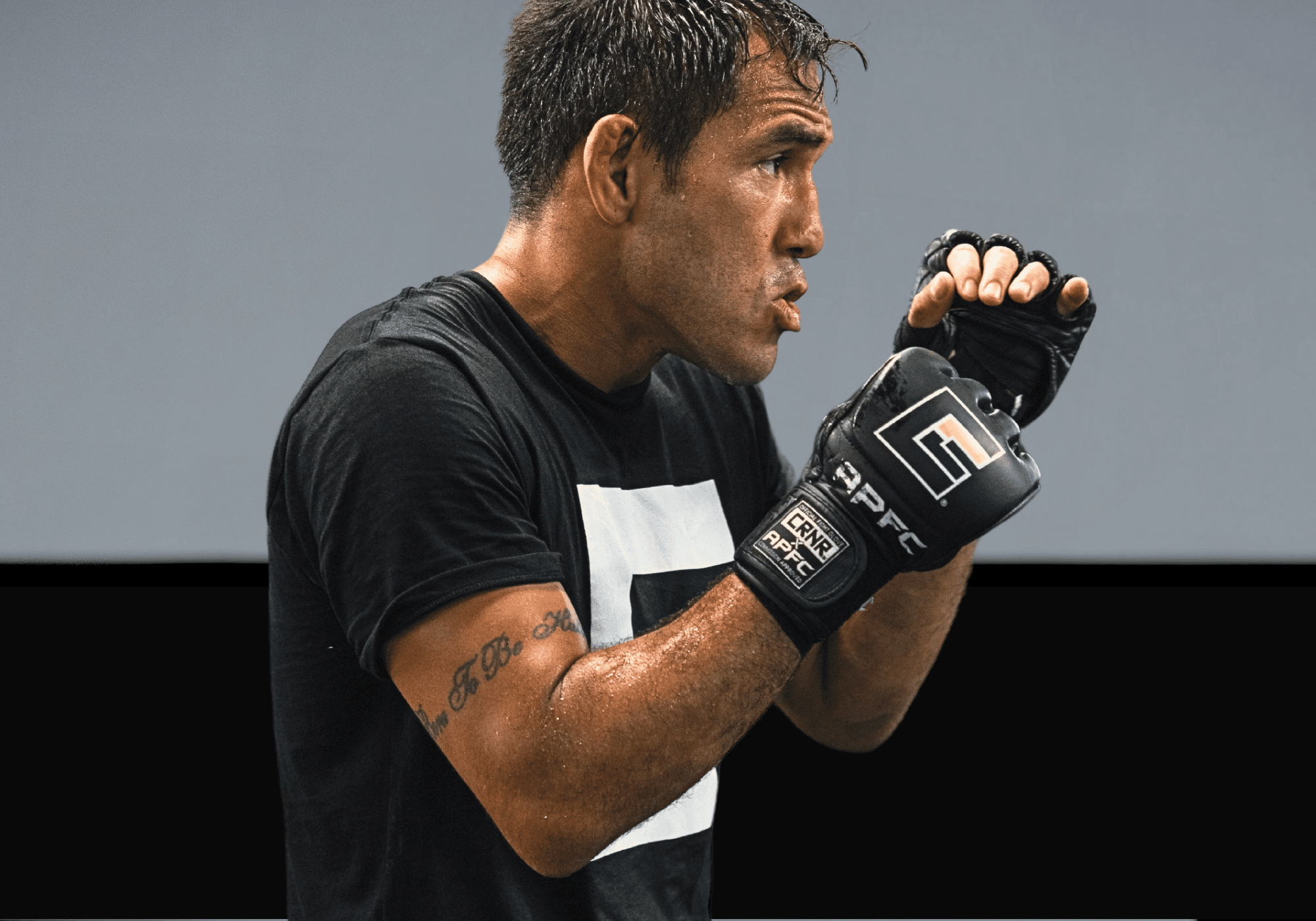 Side profile of a focused male MMA fighter in a fighting stance, wearing black Combat Corner x APFC branded open-finger MMA gloves and a black Combat Corner t-shirt.