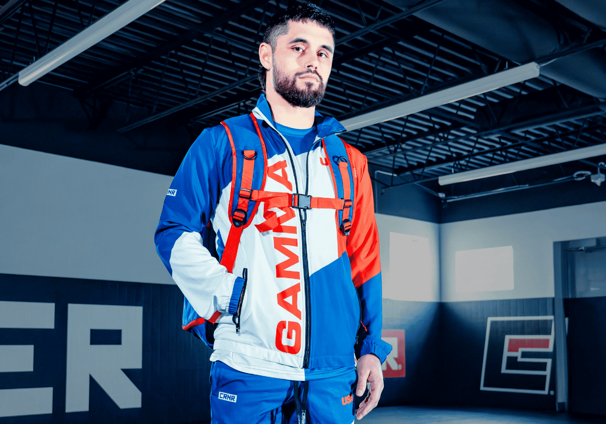 A professional studio portrait of a male athlete posing in custom Combat Corner custom GAMMA red, white, and blue team gear, including a warm-up jacket with "GAMMA" lettering, matching blue track pants, and a strapped backpack.