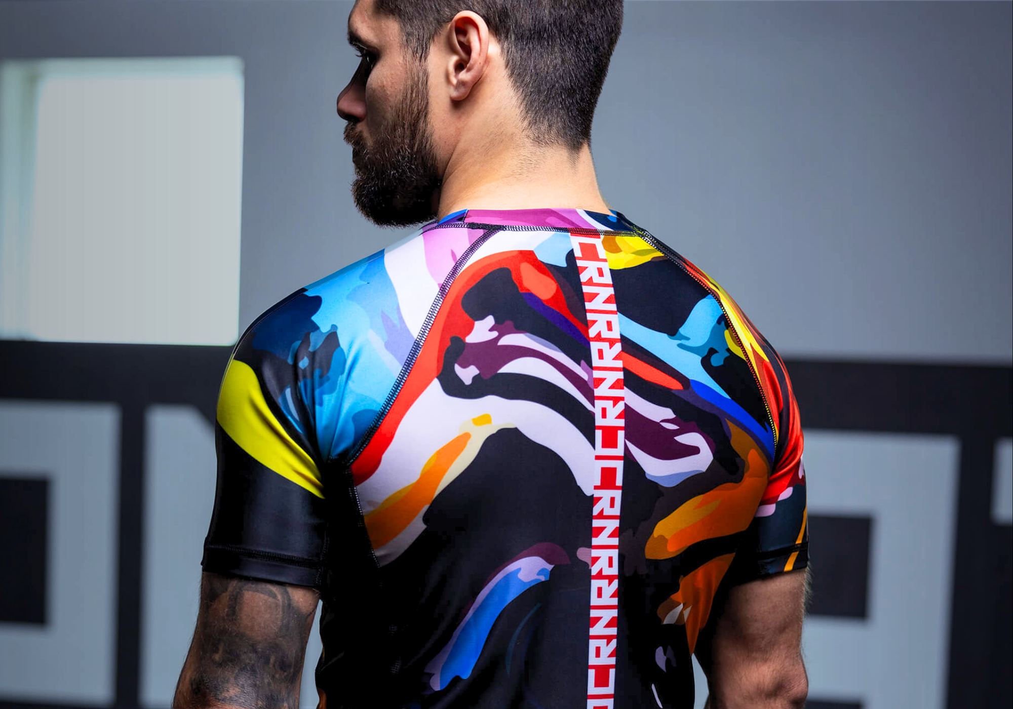 Rear view portrait of a bearded male athlete showcasing a custom Combat Corner short-sleeve rash guard with a vibrant, multicolored abstract camo pattern and a vertical spine logo.