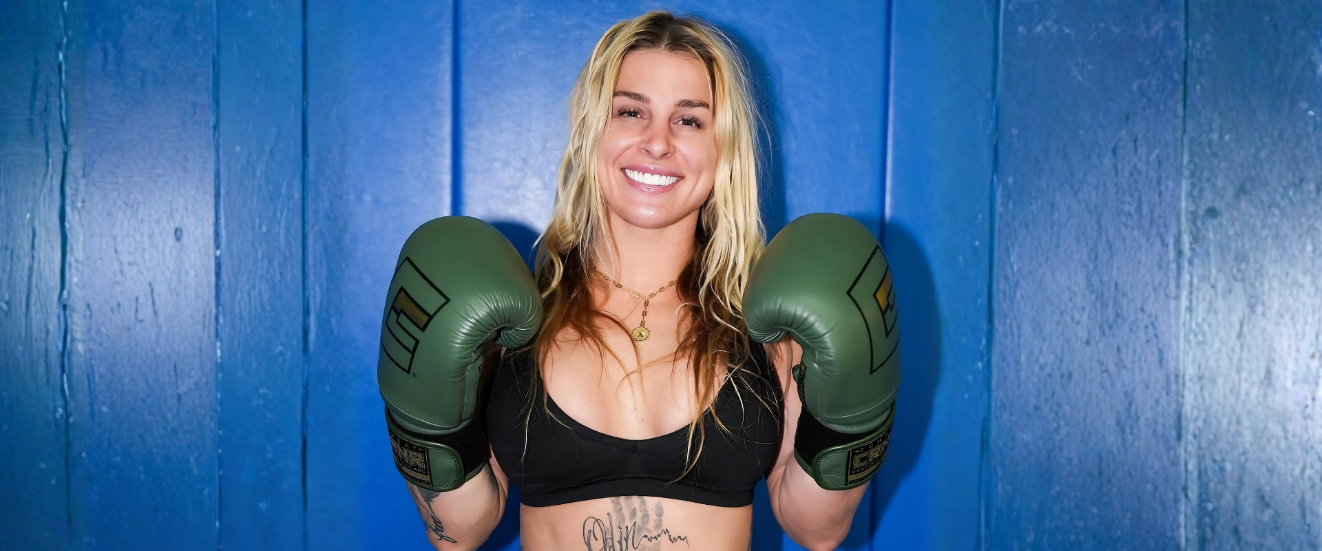 Pro fighter Hannah Goldy posing in a black sports bra and Combat Corner Boxing Gloves against a vibrant blue textured wall background. 