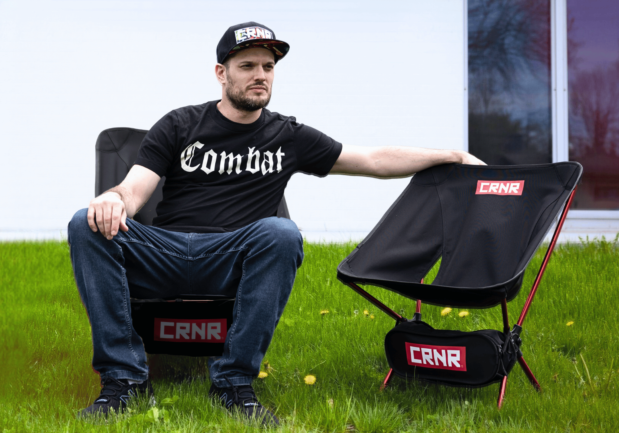 A man wearing a black Combat Corner graphic t-shirt and snapback hat sits on a grassy lawn next to a foldable black and red "CRNR" branded portable camping chair. 