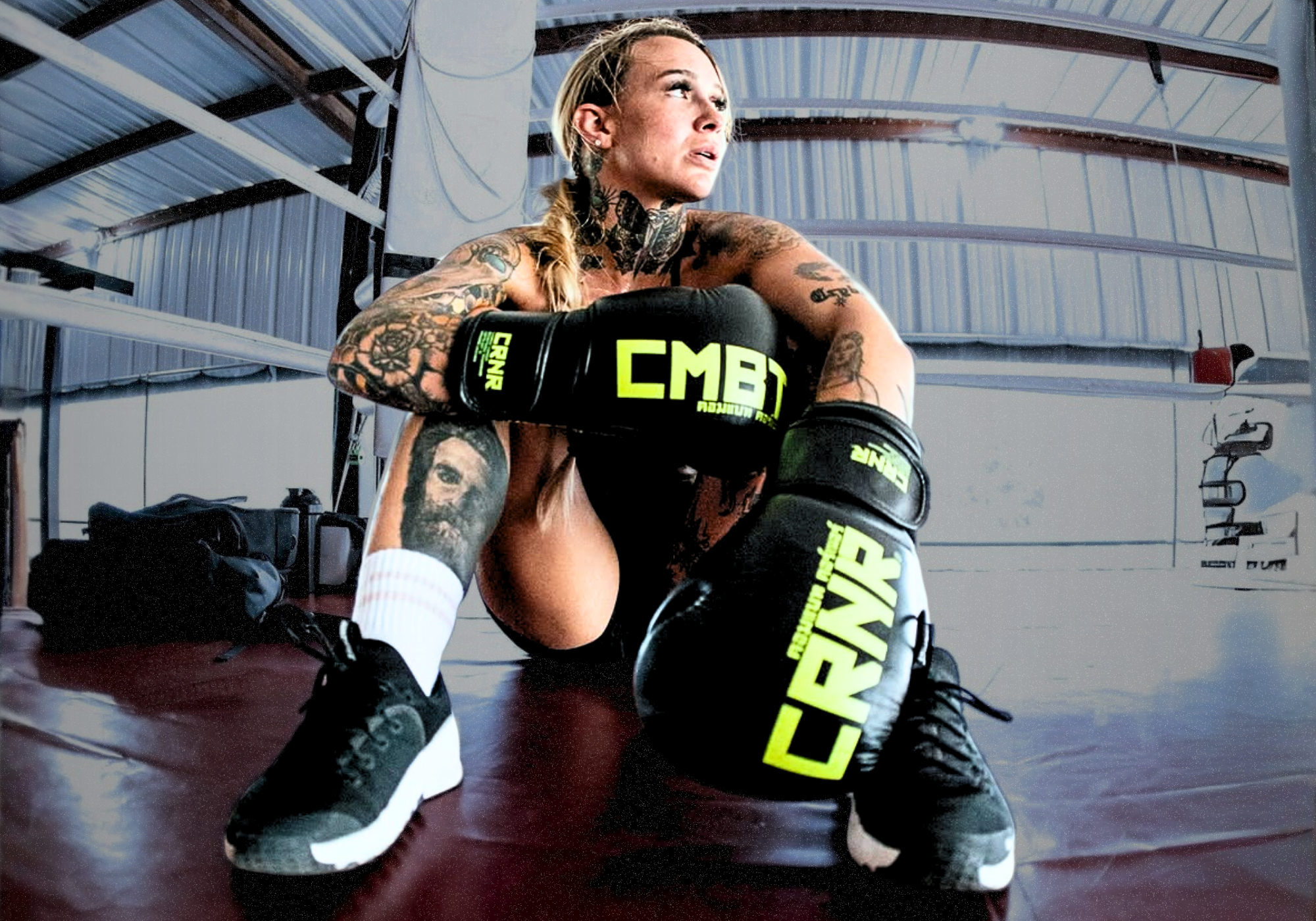 A heavily tattooed pro female fighter Taylor Starling sitting on a boxing ring mat, resting after training while wearing black Combat Corner’s chartreuse TrainAir Boxing Gloves with chartreuse/neon green and "CRNR" branding. 