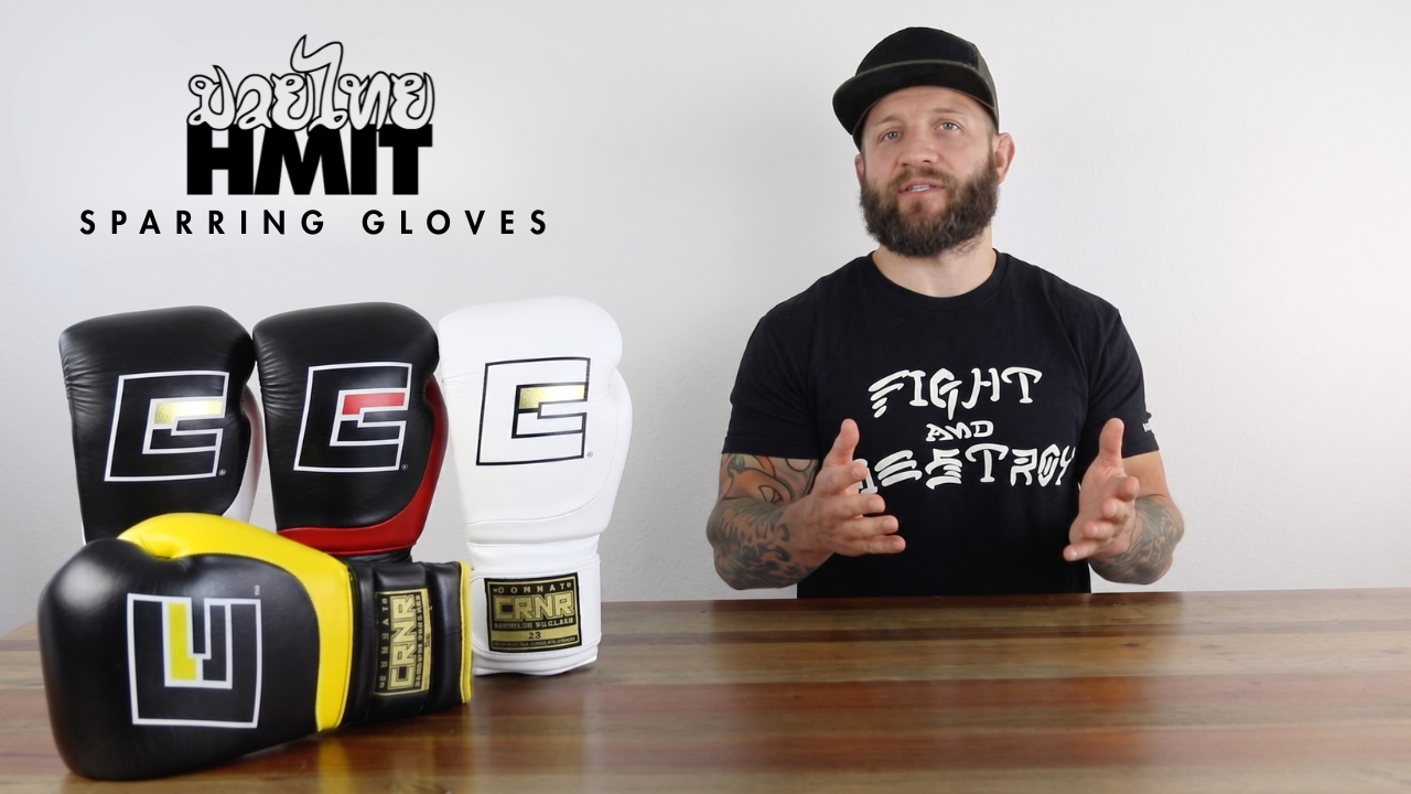 A YouTube thumbnail image featuring pro fighter Dan LaSavage giving a product walkthrough of HMIT Sparring Gloves and gesturing toward an array of colorful Combat Corner HMIT Sparring Gloves lined up on a wooden table. 