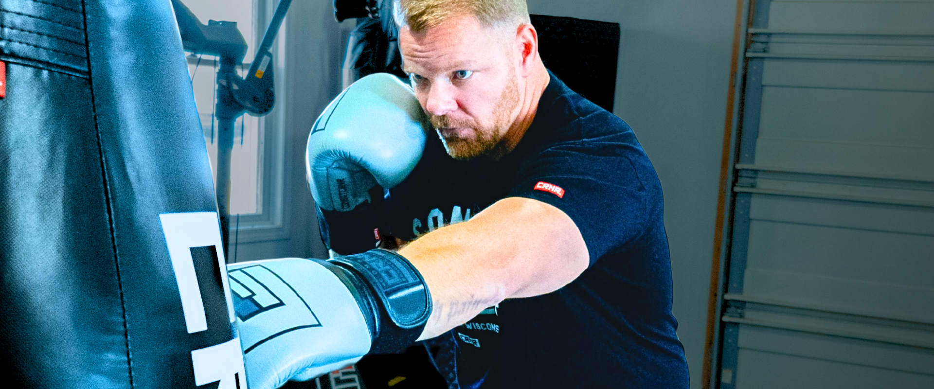 Action shot of a focused, blonde-haired male boxer training in his home gym throwing a straight punch at a Combat Corner Tear Drop heavy bag while wearing Combat Corner’s HMIT Boxing Gloves and a branded black t-shirt. 
