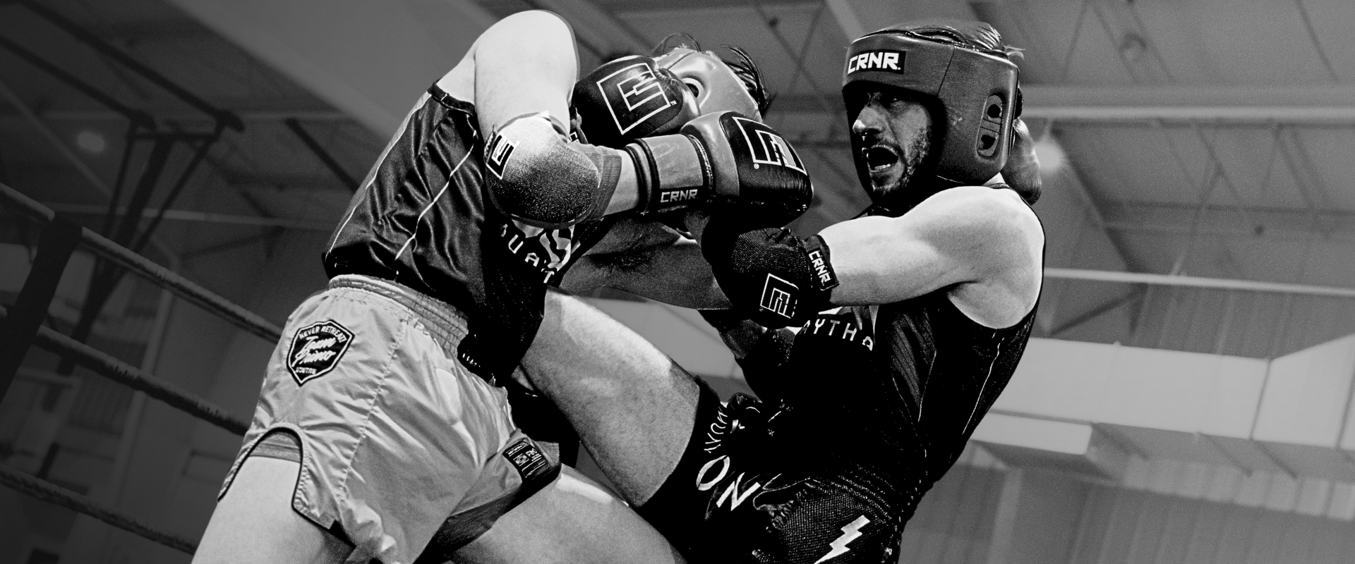 A black and white action shot of two Muay Thai fighters clinching in a ring, with one fighter throwing a knee strike while wearing Combat Corner’s Competition Headgear and matching black Combat Corner Muay Thai Elbow Pads and Slip on Shin Pads. 