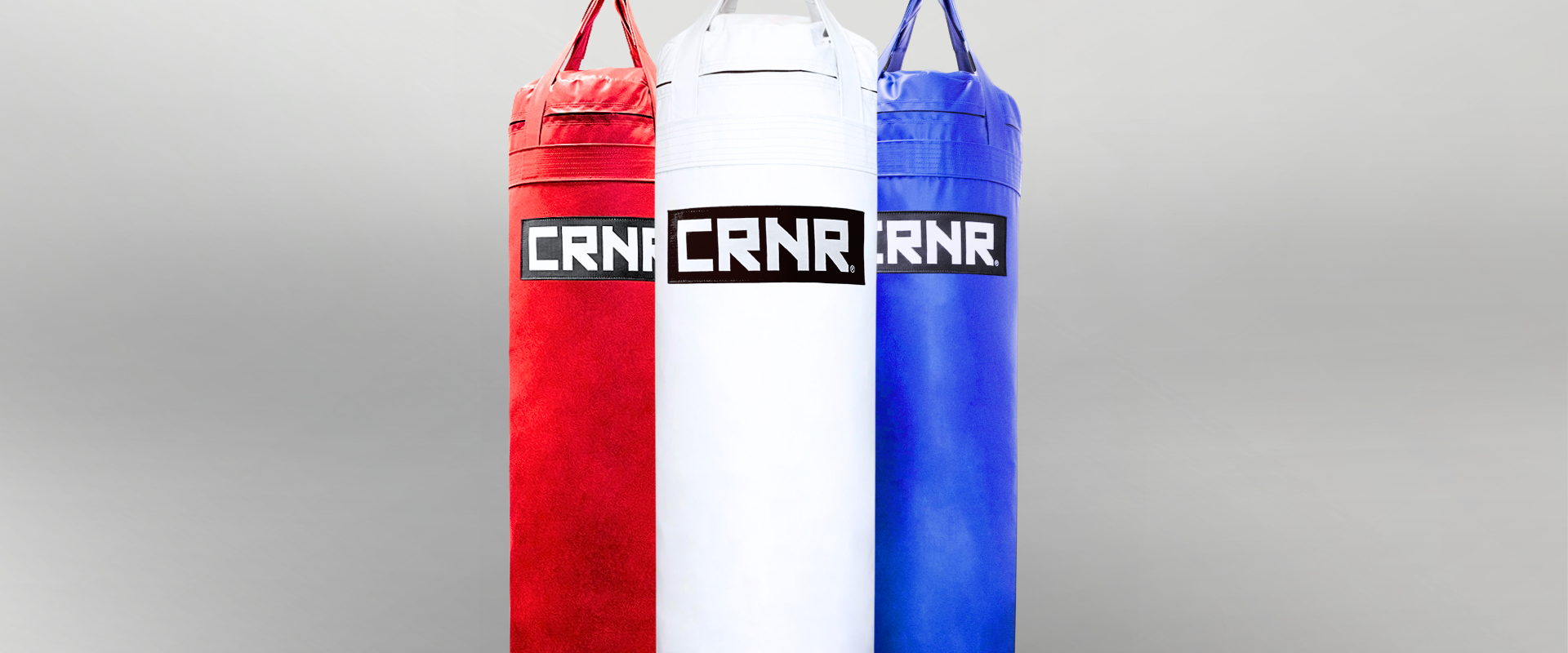 A studio shot of three Combat Corner Made in the USA heavy punching bags hanging side-by-side in red, white, and blue, each featuring a prominent black rectangle patch with the white Combat Corner "CRNR" logo. 