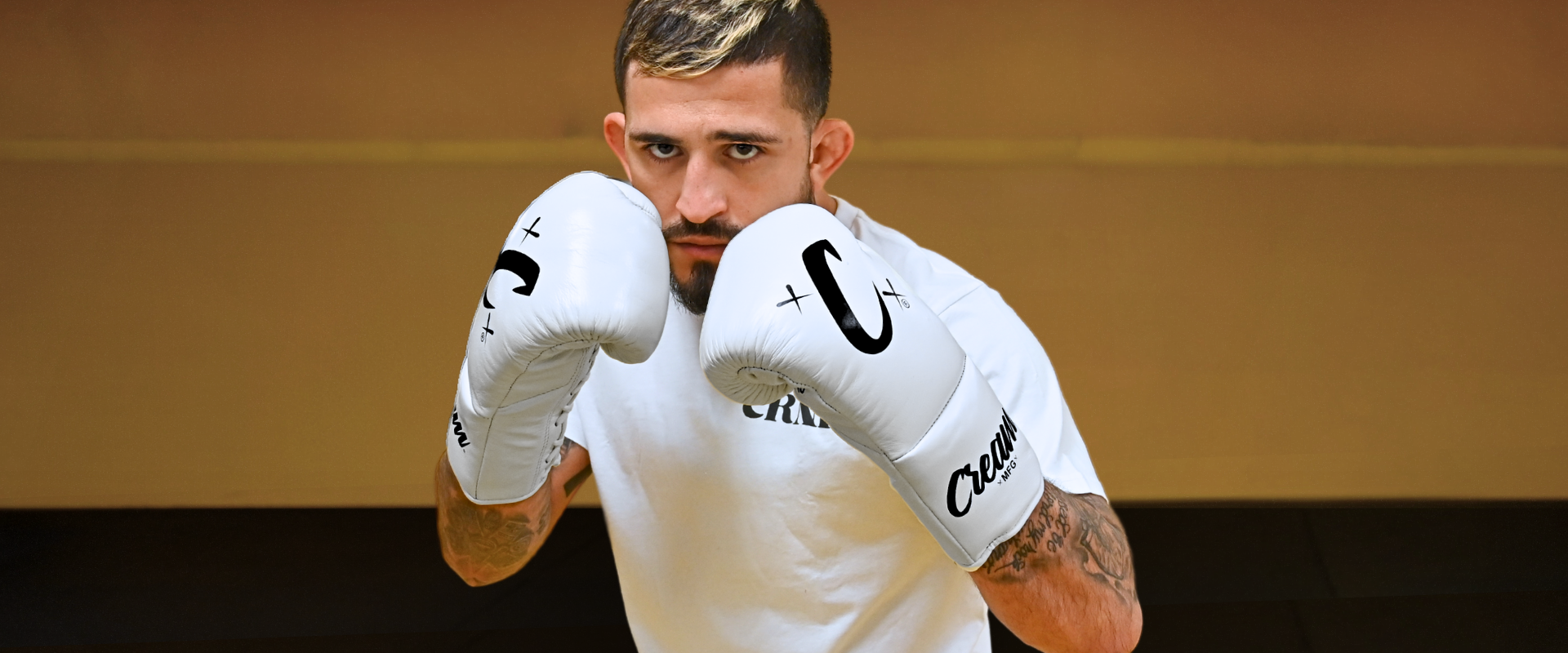Medium close-up portrait of pro fighter Sergio Pettis in a high guard stance, wearing Combat Corner’s white Relic T-Shirt and premium white leather Cream TM1 Mexican-style boxing gloves with black script branding. 