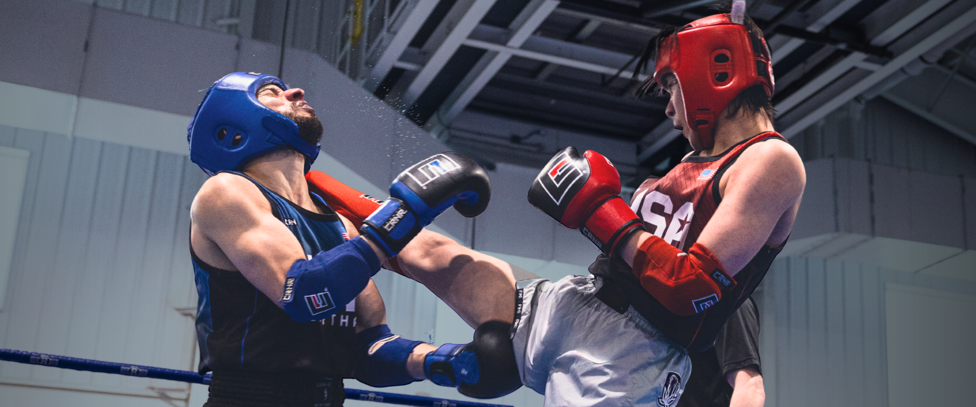 High-impact action shot of two Muay Thai fighters competing in a ring, with one fighter landing a powerful body kick while wearing official red and blue Combat Corner Muay Thai protective gear, headguards, and gloves. 
