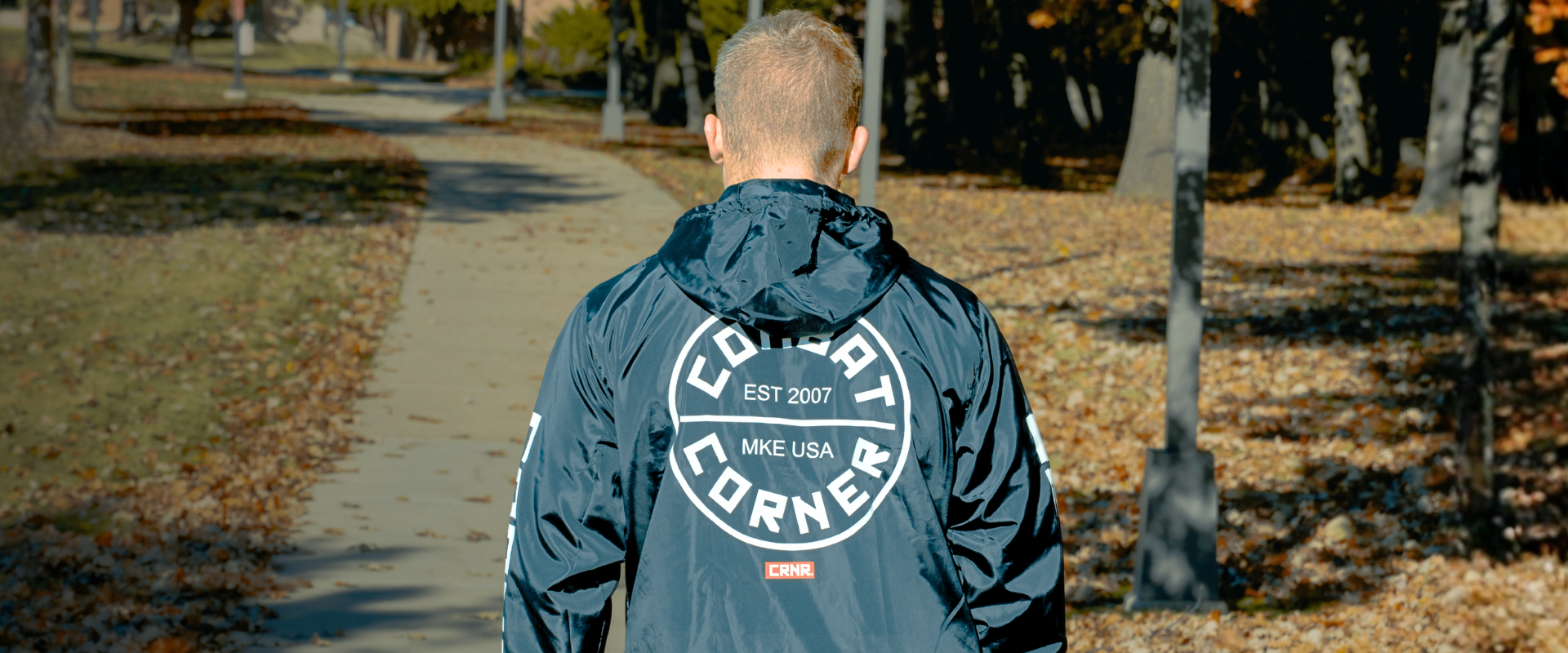 Medium shot of a blonde-haired man with his back to the camera wearing Combat Corner’s WindBreaker jacket with a square "CRNR" chest logo and bold sleeve text. 
