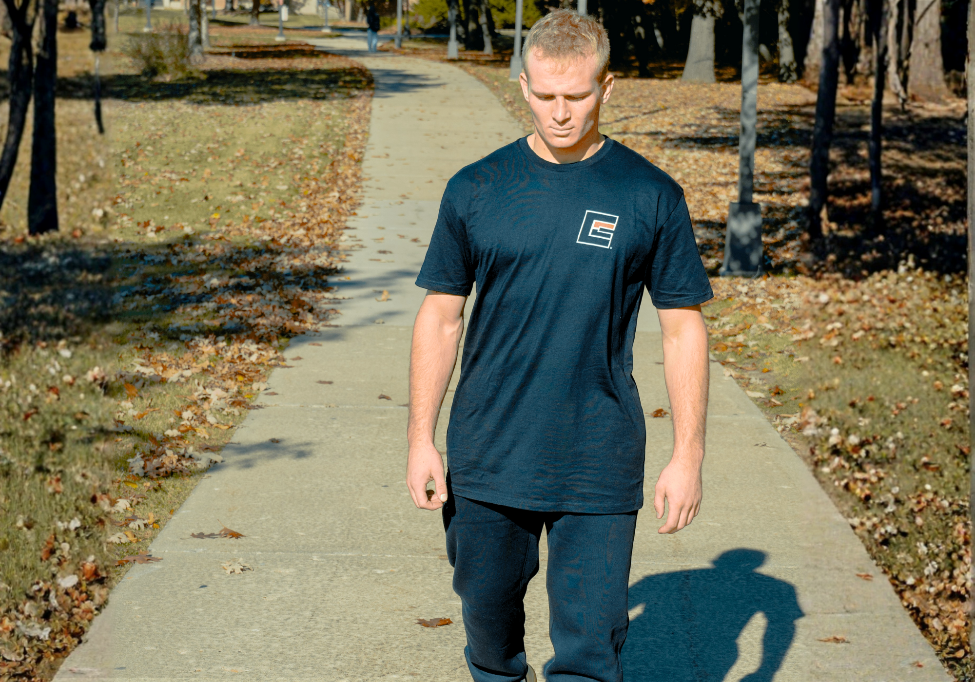 A blonde-haired man walking down a park path, looking downward while wearing Combat Corner’s Fight Equip T-Shirt featuring a small square chest logo and Combat Corner’s Relaxed Sweatpants. 