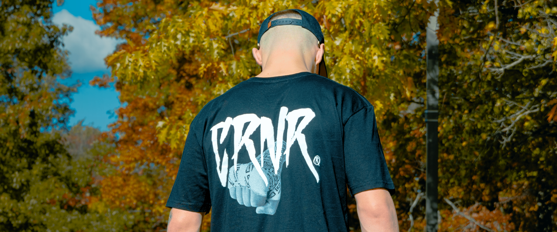 Rear view of a man wearing a black Combat Corner t-shirt featuring a large white, stylized "CRNR" graffiti-style logo and a graphic of a wrapped fist, set against a background of autumn trees.