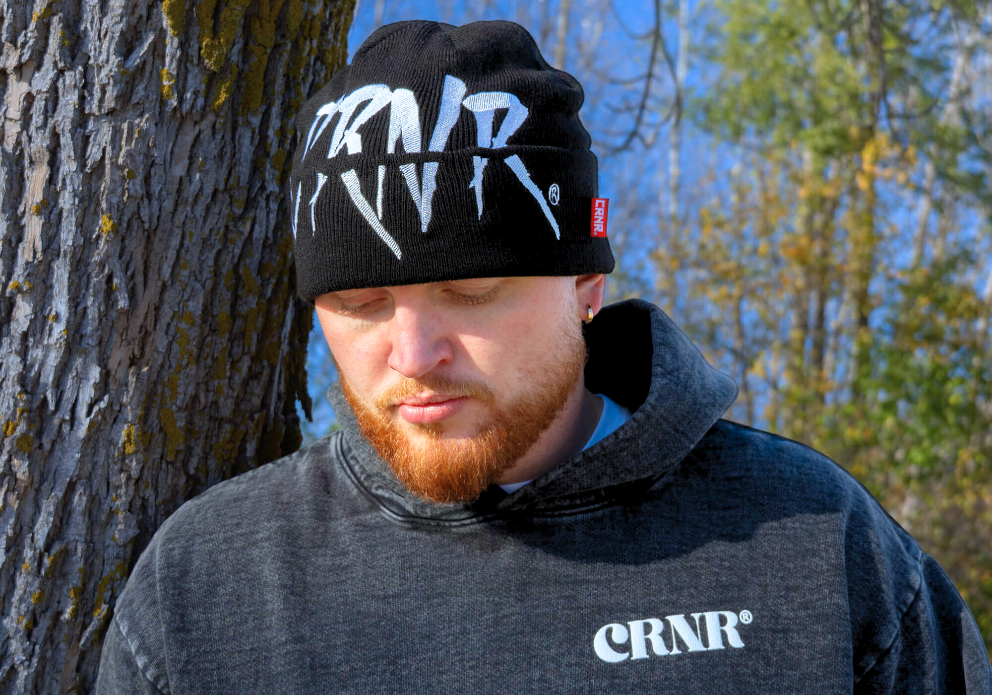 Medium close-up portrait of a red-bearded man leaning against a tree, wearing Combat Corner’s Relic Hoodie and Shred Beanie.