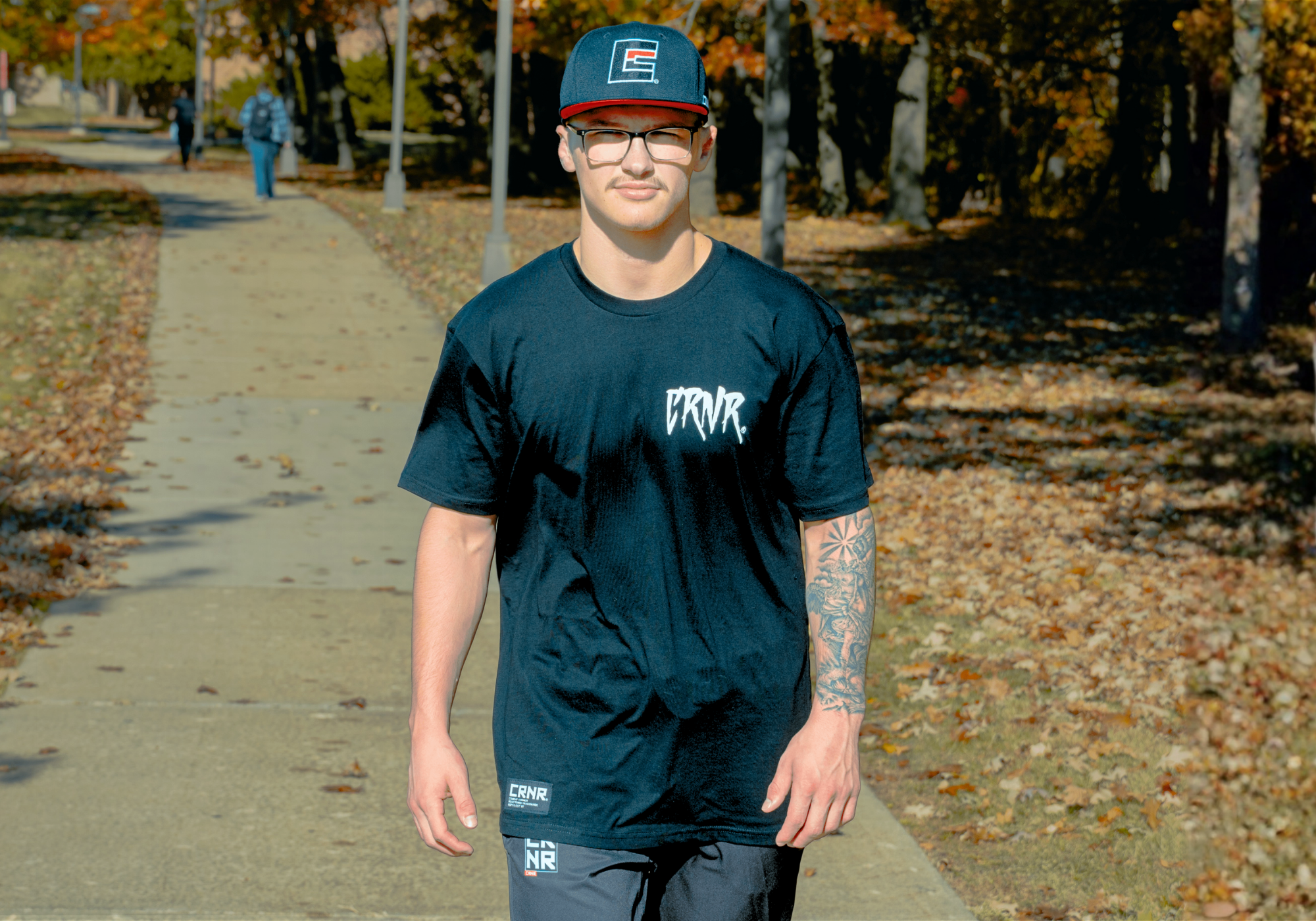 A young man with glasses, a mustache, and a tattooed arm walking down an outdoor park path, wearing Combat Corner’s Icon Snapback Hat, That’s A Wrap T-Shirt with a white stylized logo and CRNR Tech Joggers. 