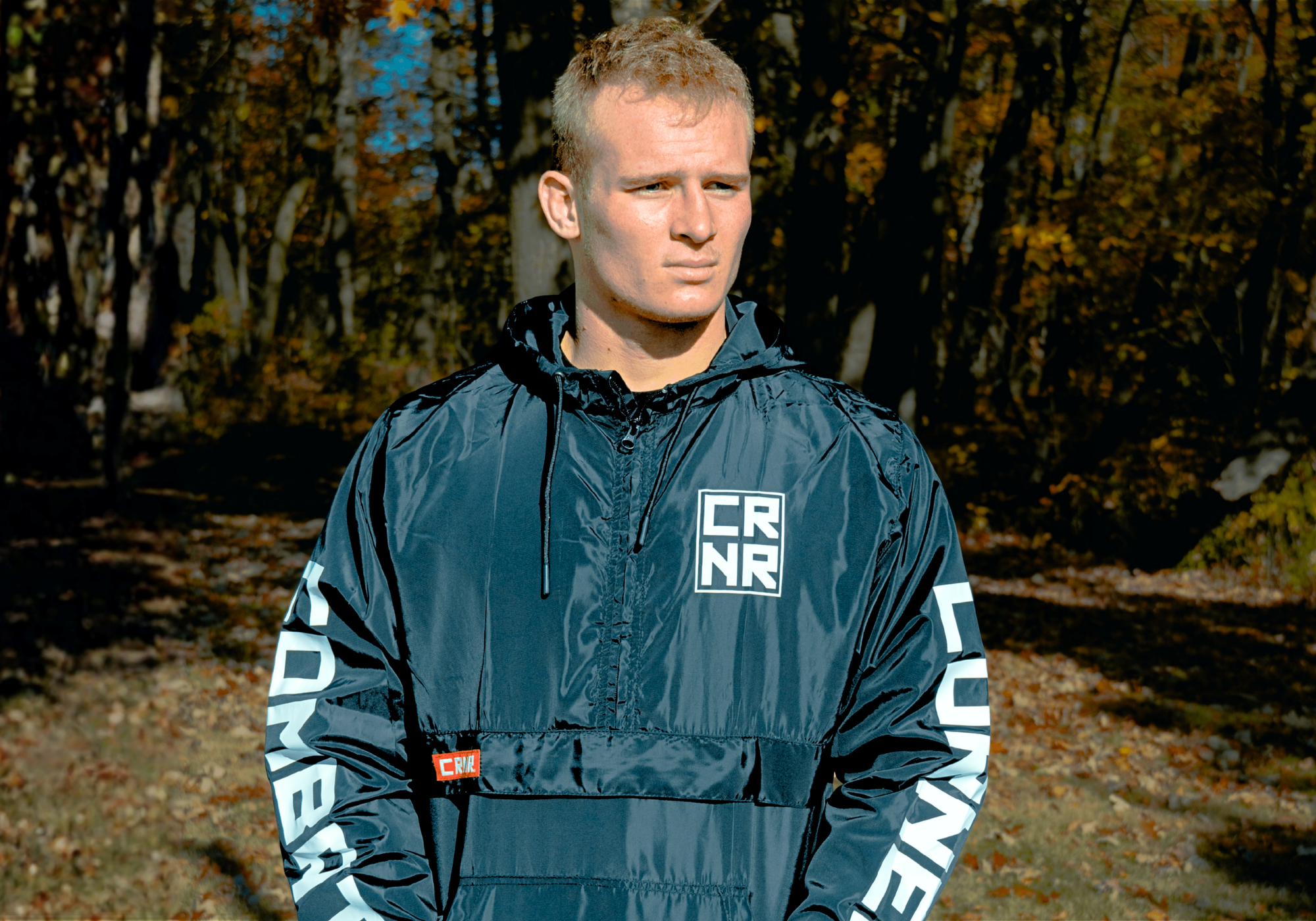 Medium shot of a blonde-haired man looking to the side outdoors, wearing Combat Corner’s WindBreaker jacket with a square "CRNR" chest logo and bold sleeve text. 