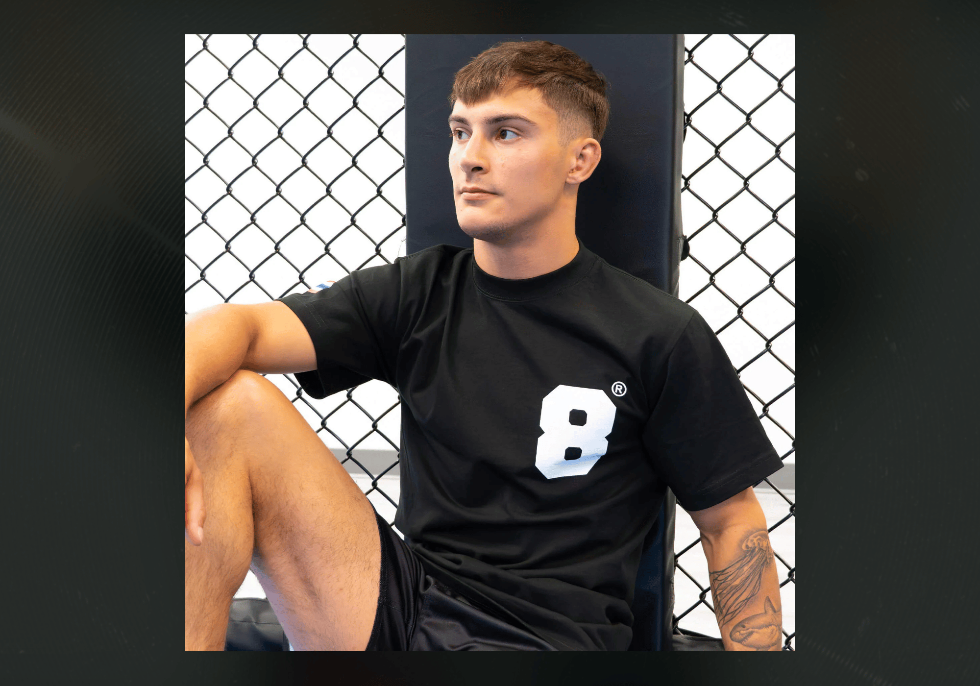 A male athlete sitting against a padded corner post inside an MMA cage, wearing the black Combat Corner 8 Thai Boxing T-Shirt featuring a large white number "8" printed on the chest.