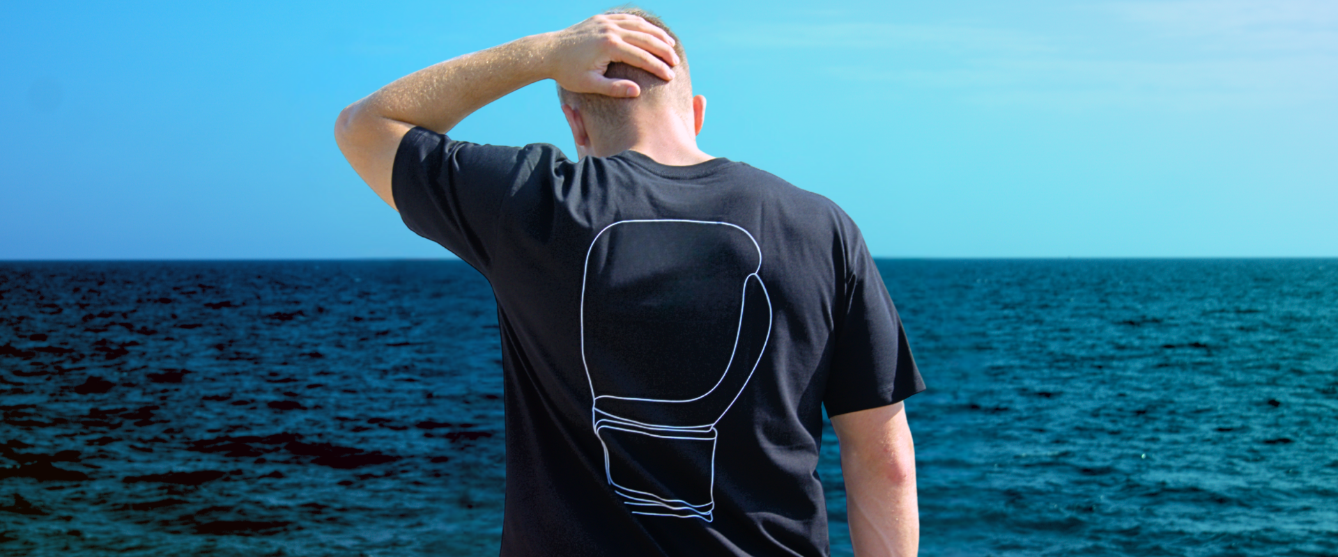 Portrait of a blonde-haired man with his back to the camera looking into the distance against a bright blue sky, wearing Combat Corner’s HMITrace T-Shirt. 