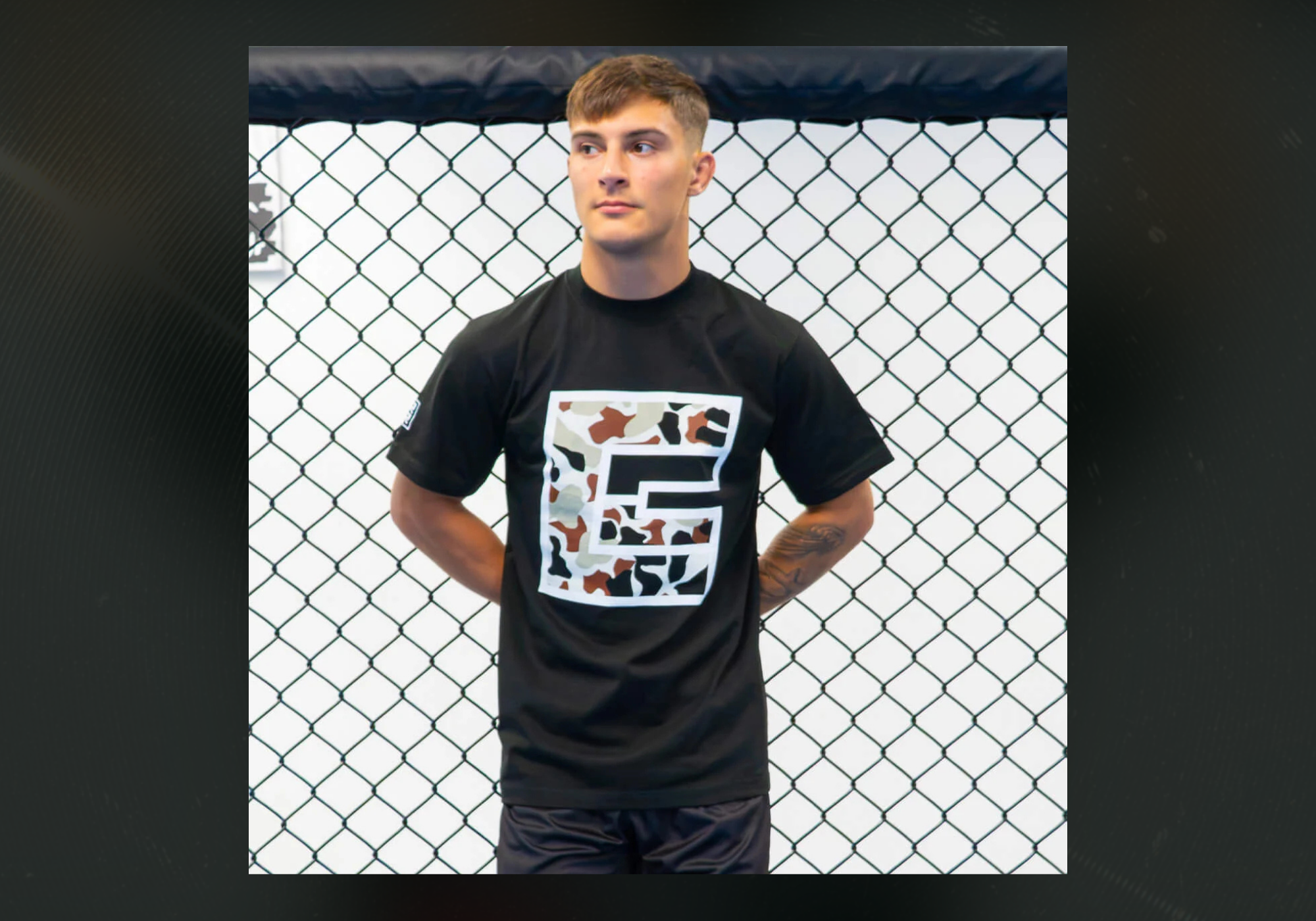 Front view of an athlete standing inside a chain-link fence cage, wearing the black Combat Corner Camo Icon T-Shirt featuring a large white square logo filled with a camouflage pattern.