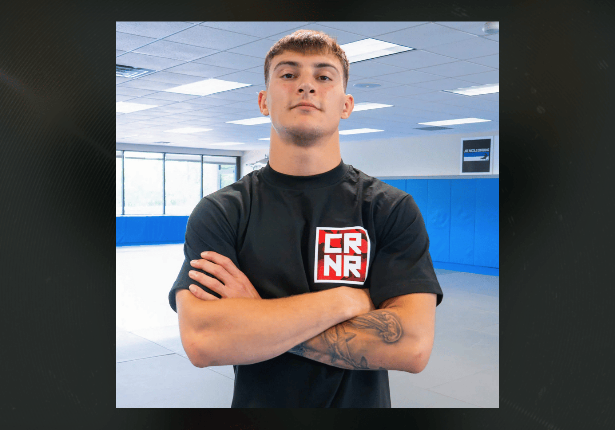 A front portrait of a young male athlete standing with arms crossed in a gym, wearing the black Combat Corner Cinder Topo Stacked T-Shirt featuring a red camouflage square logo with white "CRNR" lettering.