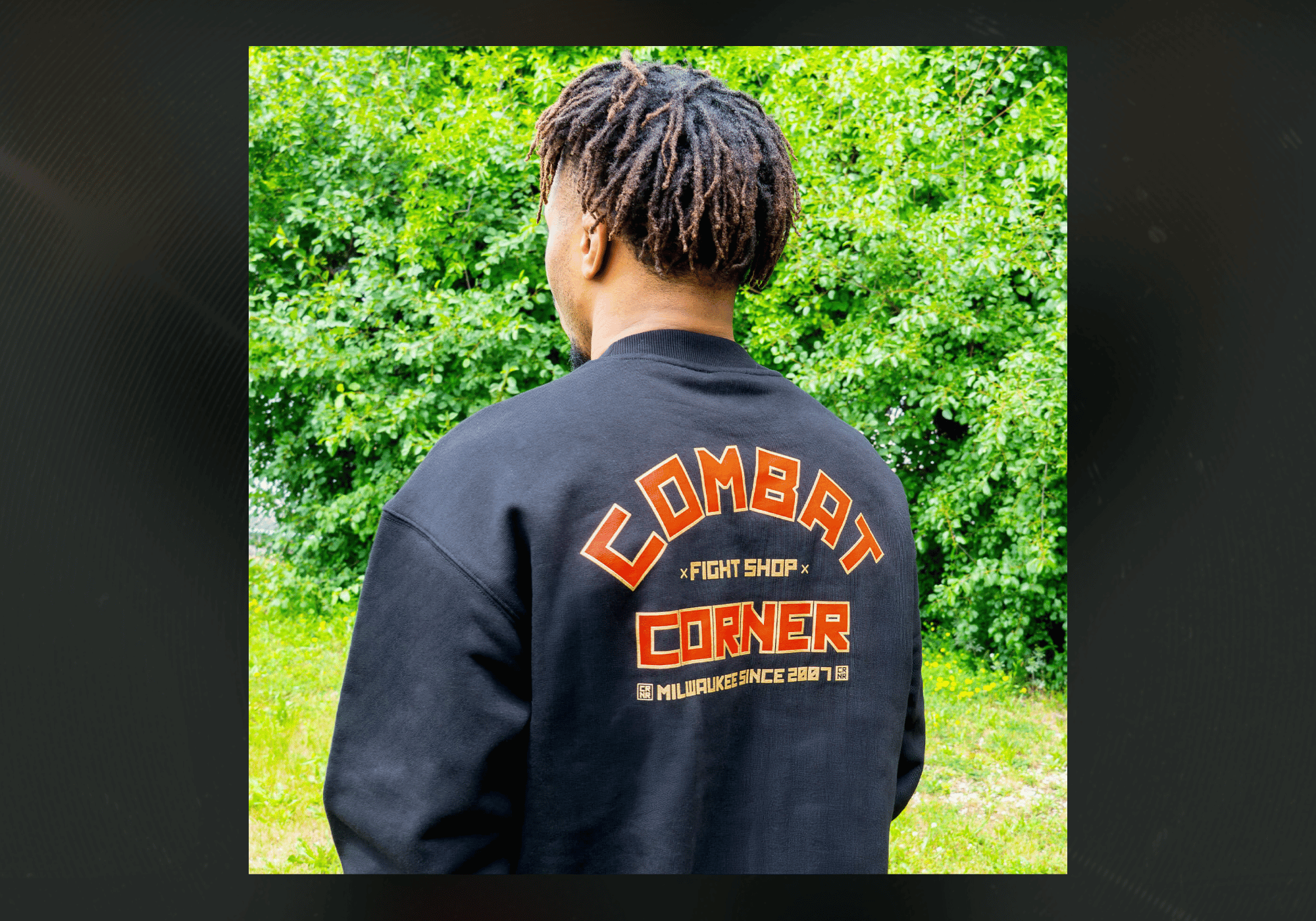 Rear view of a man with dreadlocks standing outdoors, wearing the black Combat Corner Fire Fight Crewneck Sweatshirt featuring a large orange, red, and gold "Combat Corner Fight Shop" graphic printed on the back.