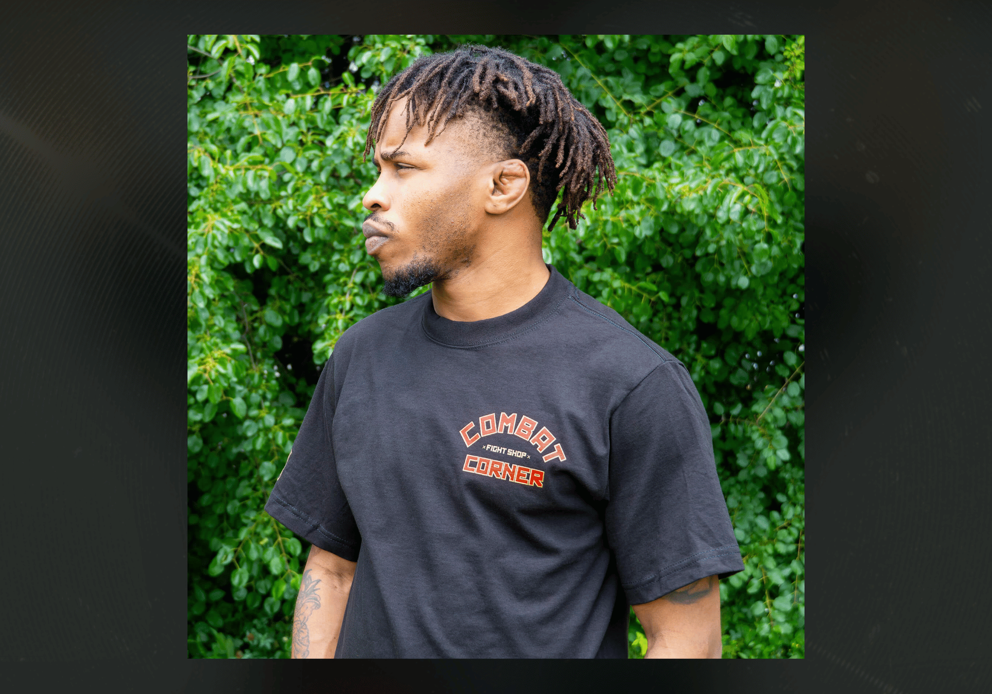 A three-quarter profile shot of a man with dreadlocks standing outdoors, wearing the black Combat Corner Fire Fight T-Shirt with an arched orange and red "Combat Corner Fight Shop" logo on the left chest.