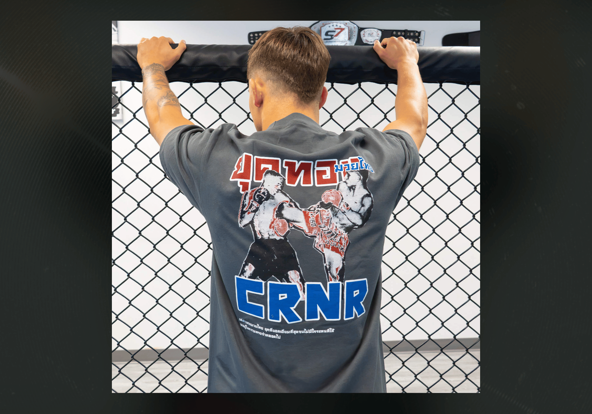 Rear view of an athlete leaning against a chain-link cage in a gym, wearing the grey Combat Corner Golden Era Thai Boxing T-Shirt featuring a vintage Muay Thai fighter illustration with "CRNR" lettering and Thai script.
