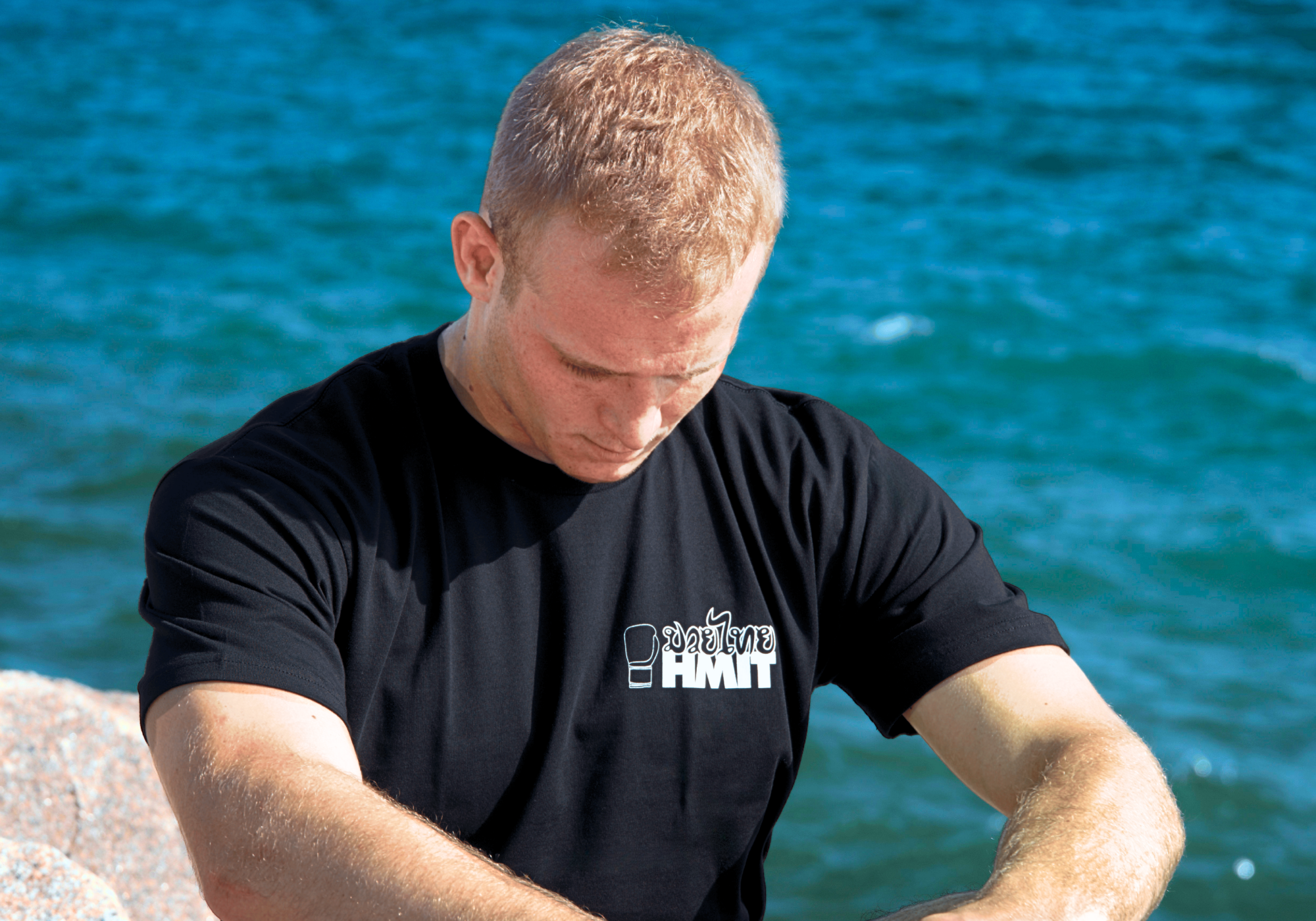 Portrait of a blonde-haired man looking down against a bright blue sky, wearing Combat Corner’s HMITrace T-Shirt. 