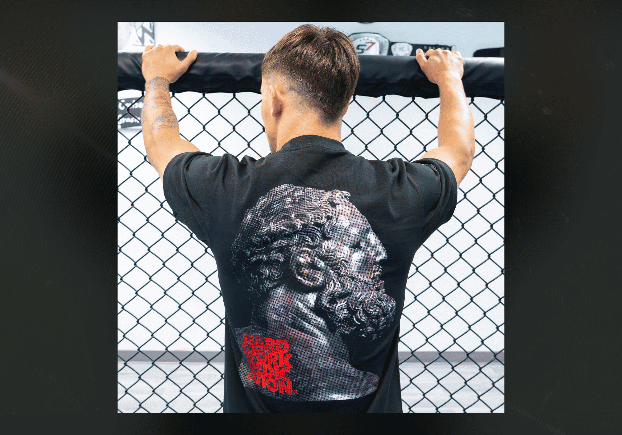 Rear view of an athlete leaning against a chain-link cage in a gym, wearing the black Combat Corner HWD Ancient T-Shirt featuring a detailed metallic print of an ancient philosopher's statue bust with red text.