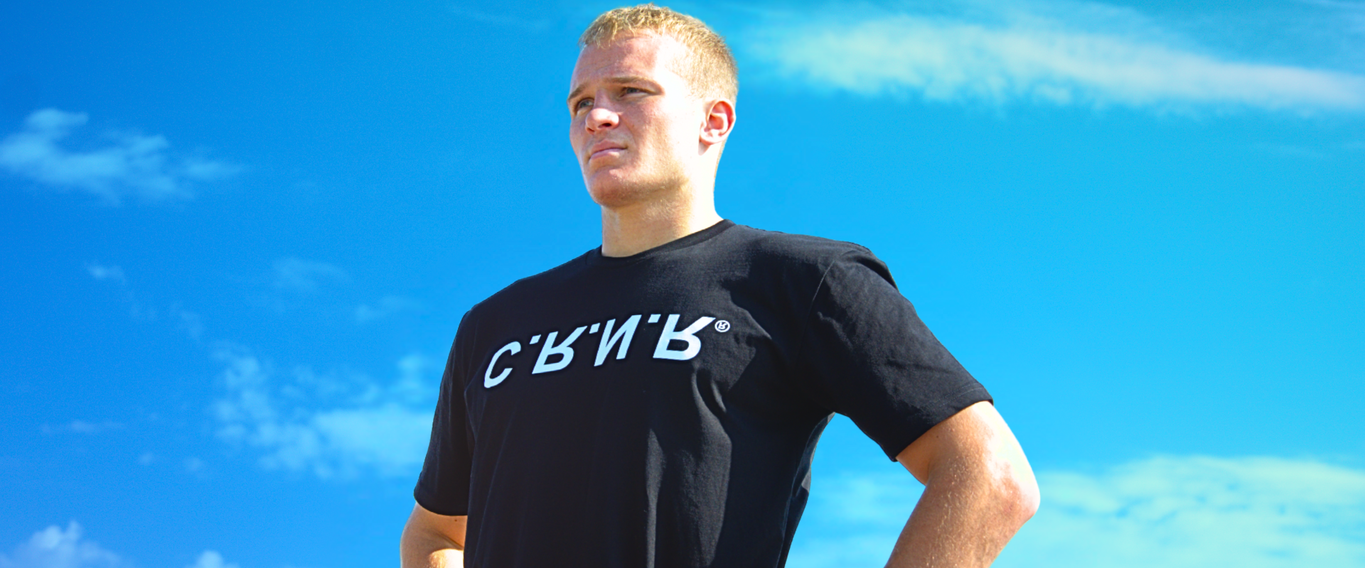 Low-angle portrait of a blonde-haired man looking into the distance against a bright blue sky, wearing Combat Corner’s UP DN IN OUT T-Shirt featuring a stylized white "CRNR" logo across the chest.