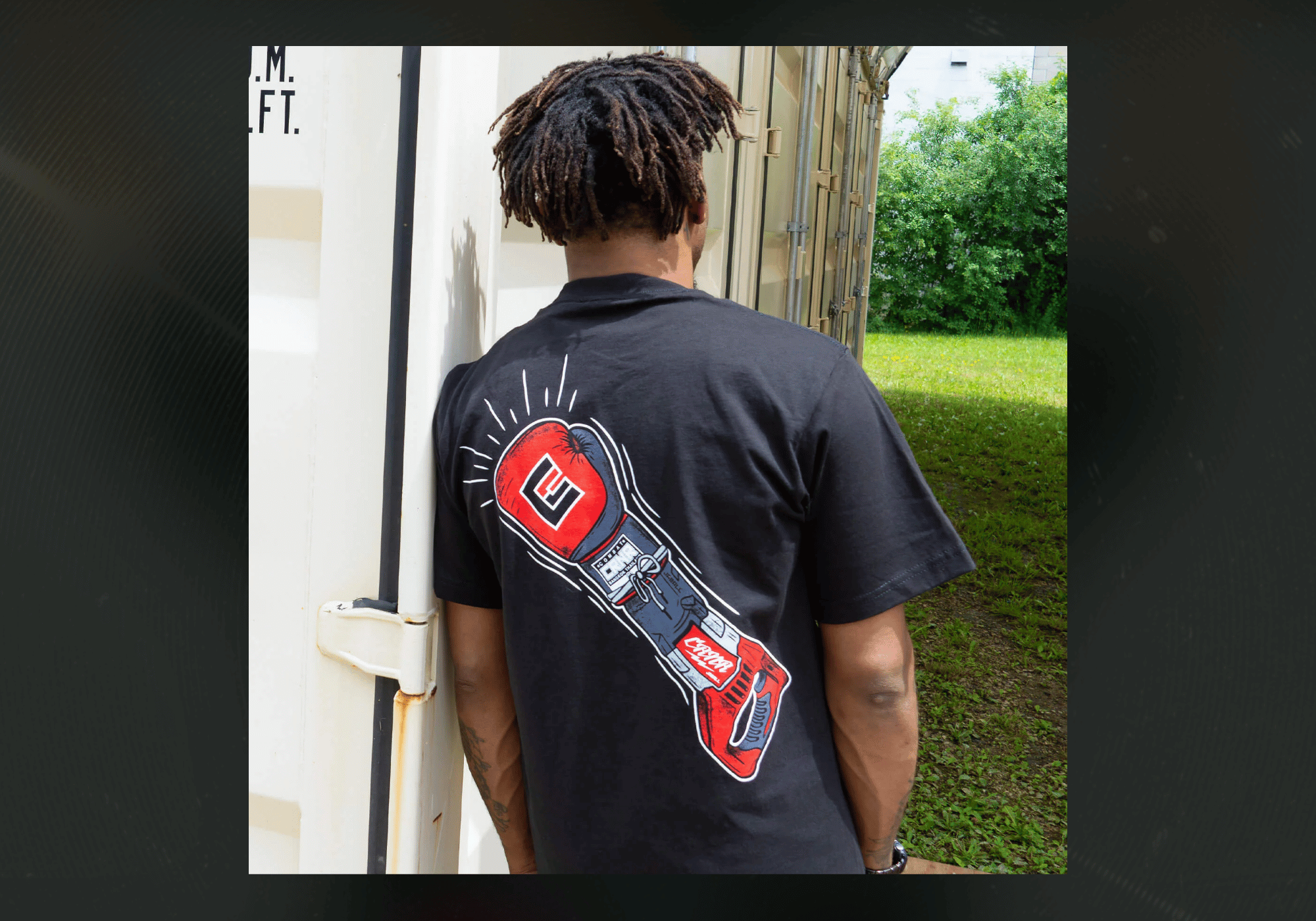 Rear view of a man leaning against a white shipping container, wearing the black Combat Corner JabAll T-Shirt featuring a stylized illustration of a red boxing glove merged with a power saw handle.
