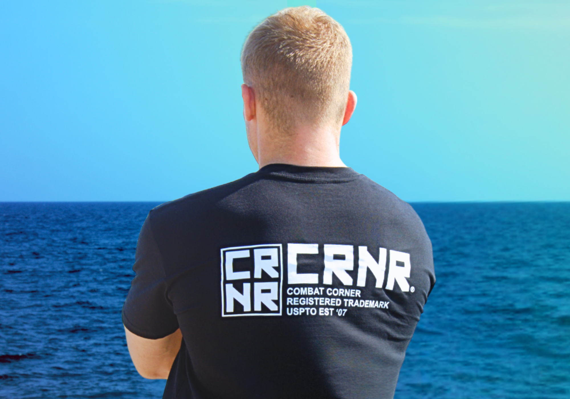 Portrait of a blonde-haired man with his back to the camera looking into the distance against a bright blue sky, wearing Combat Corner’s Stacked T-Shirt.