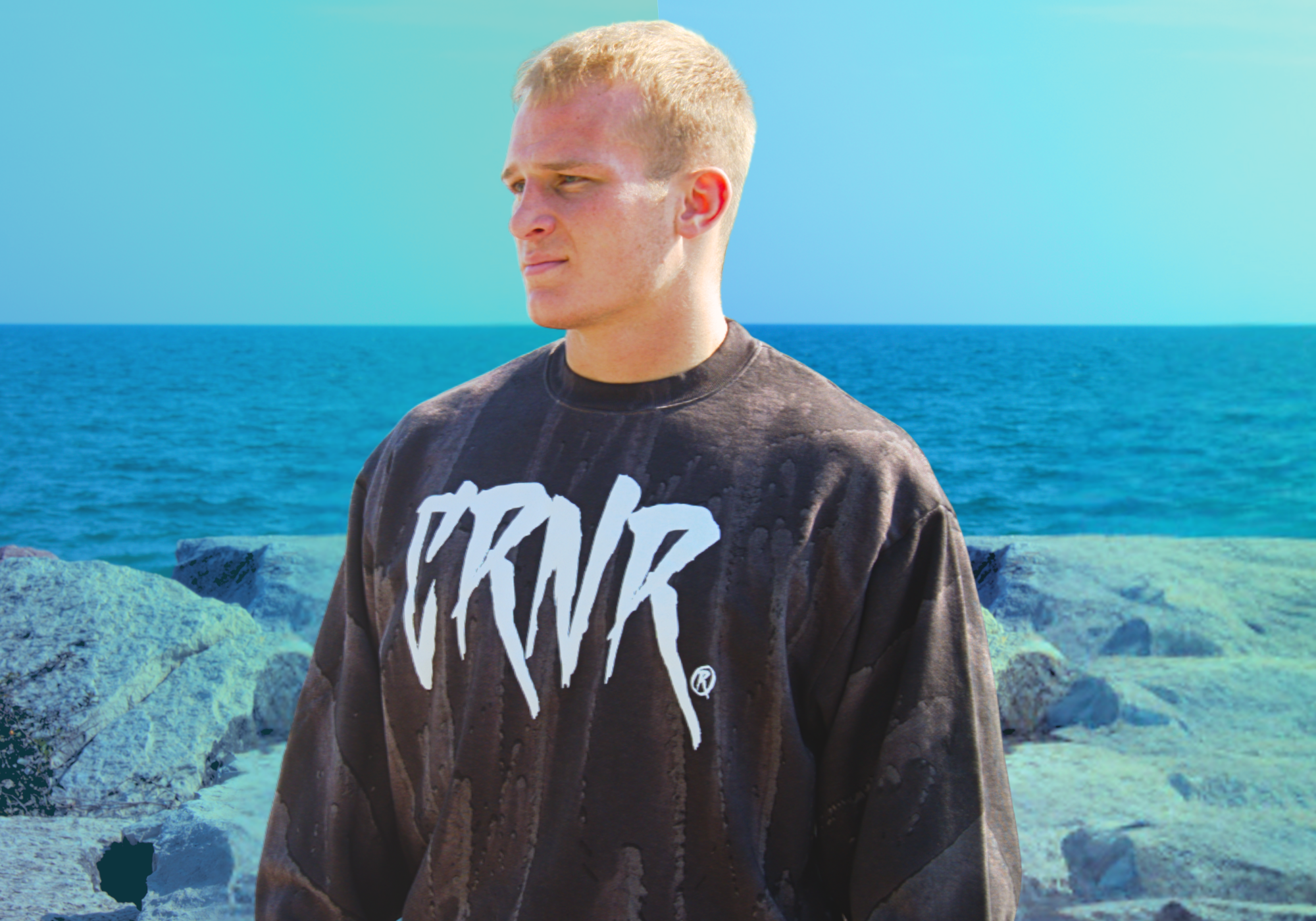 Low-angle portrait of a blonde-haired man looking into the distance against a bright blue sky, wearing Combat Corner’s That's A Wrap Crewneck sweater.