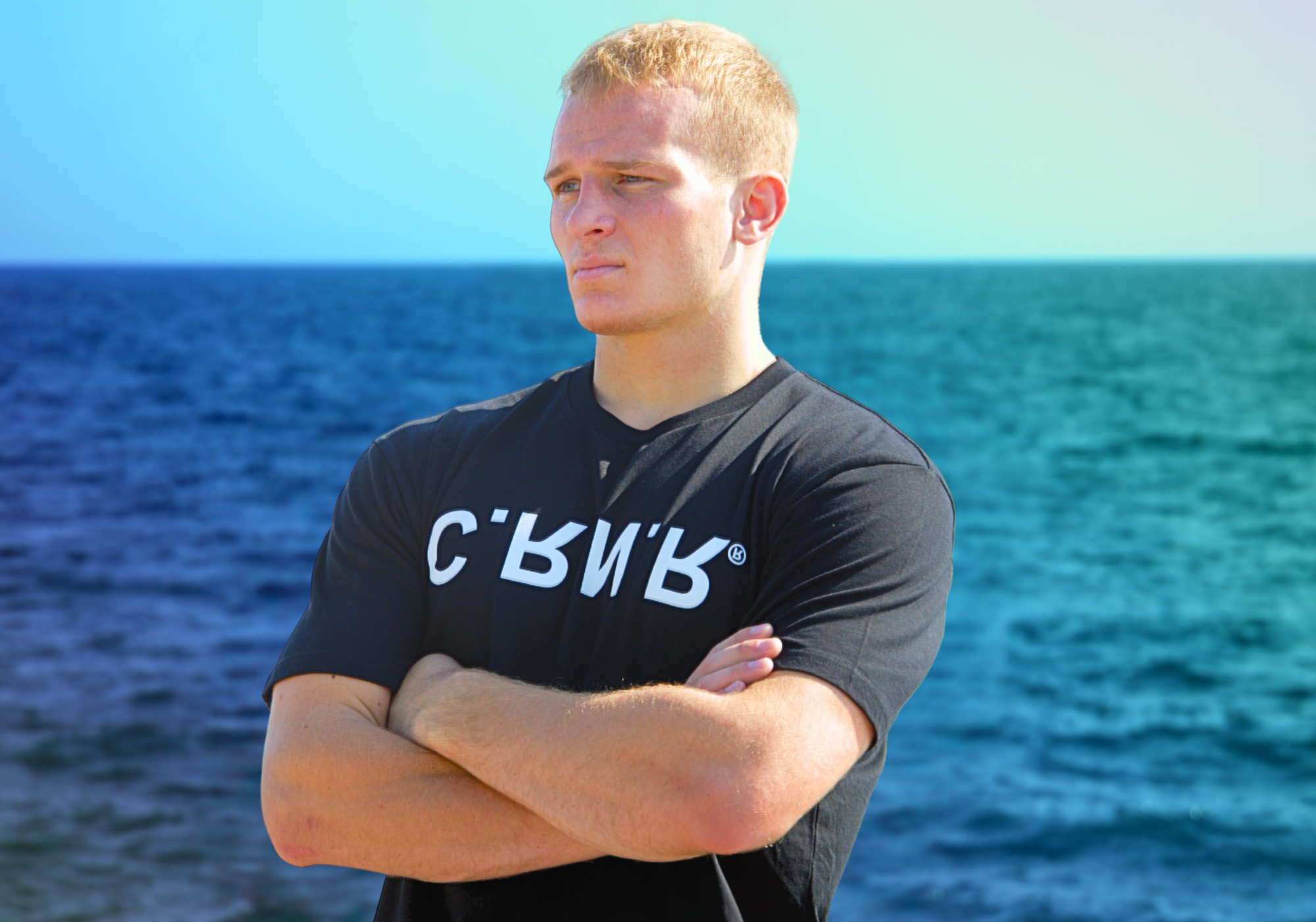 Portrait of a blonde-haired man looking into the distance against a bright blue sky, wearing Combat Corner’s UP DN IN OUT T-Shirt featuring a stylized white "CRNR" logo across the chest.