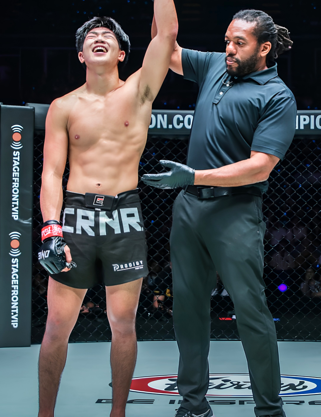 Inside a ONE Championship cage, referee Herb Dean raises the arm of a ONE World Champion and Combat Corner Pro Team Member Adrian Lee who is wearing black Combat Corner’s black Supreme Hybrid Fight Shorts with a large grey "CRNR" logo and matching black "ONE" fight gloves. 