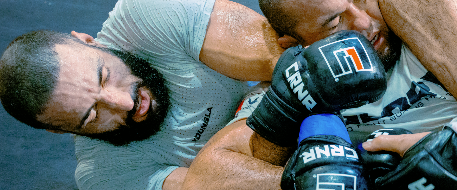 An intense close-up action shot of professional fighter Belal Muhammad grappling on the mat during training, while his partner defends wearing Combat Corner’s black Prime MMA Sparring Gloves with prominent white and red "CRNR" logos.