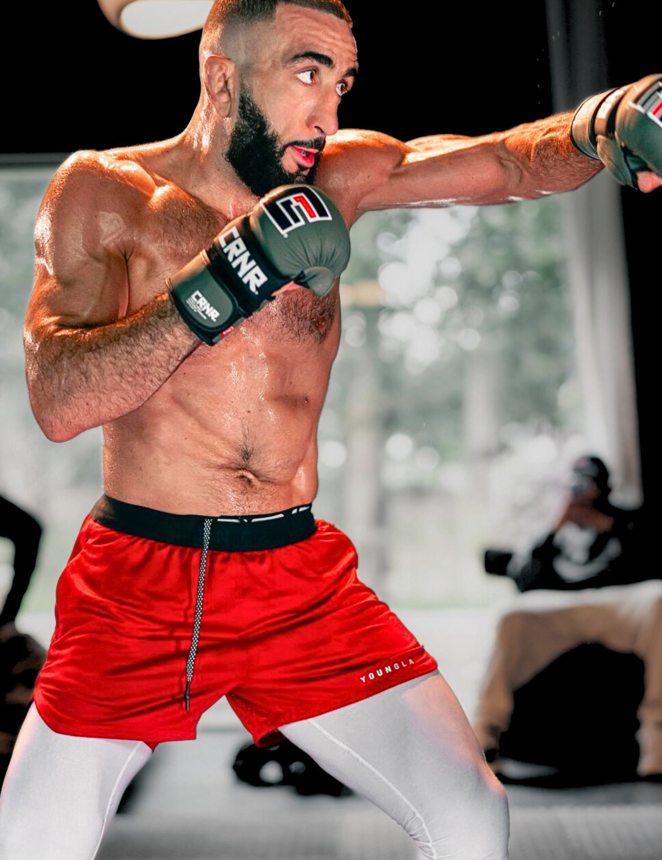 An action shot of UFC World Champion and Combat Corner Pro Team Member Belal Muhammad training with Combat Corner’s OD green MMA Spar Gloves. He is also wearing bright red shorts over white compression leggings. 