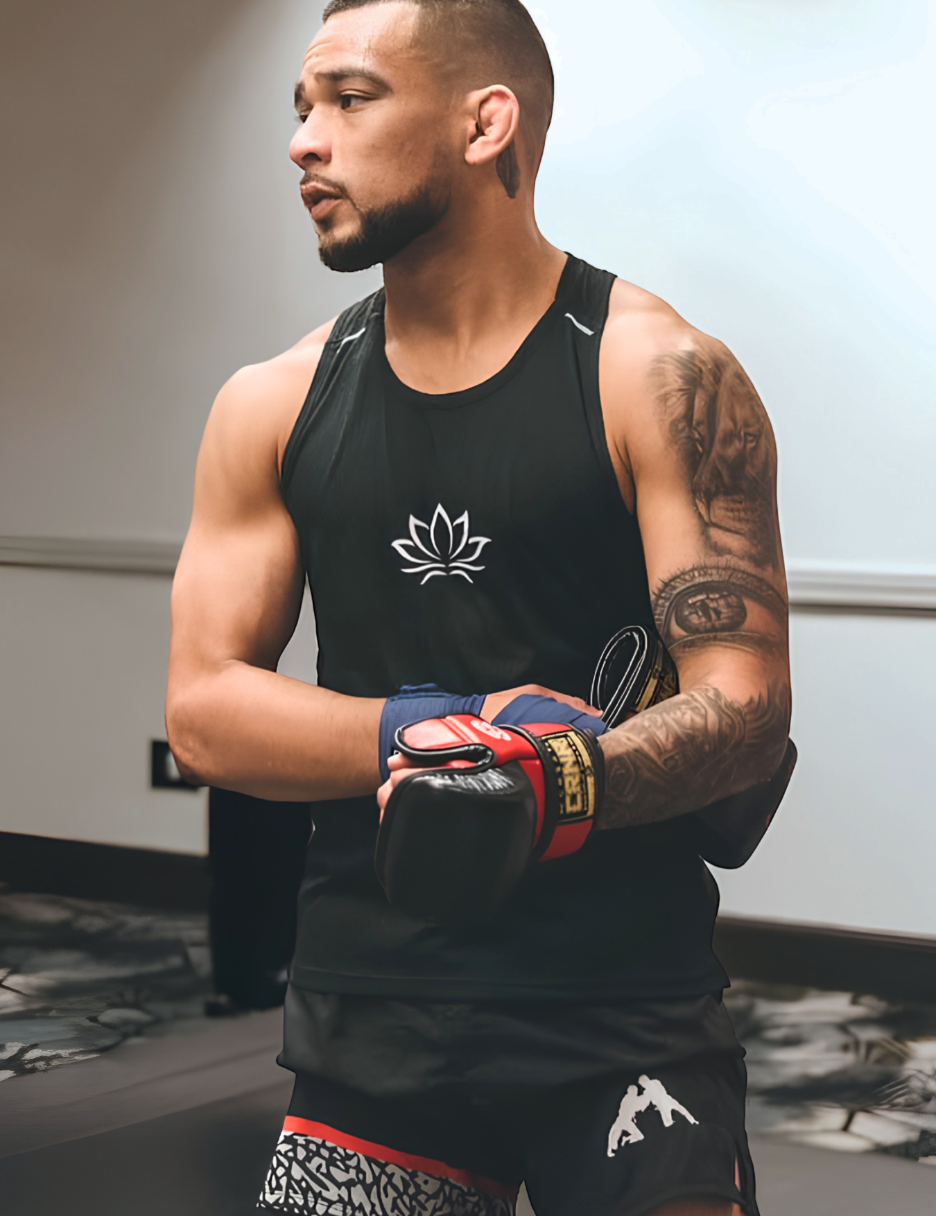Three-quarter portrait of UFC Fighter and Combat Corner Pro Team Member Christian Rodriguez with a tattooed sleeve looking to the side, wearing a black tank top with a lotus logo, Combat Corner’s black BJJ Life fight shorts, blue hand wraps, and Combat Corner’s red and black HMIT MMA Training Gloves. 