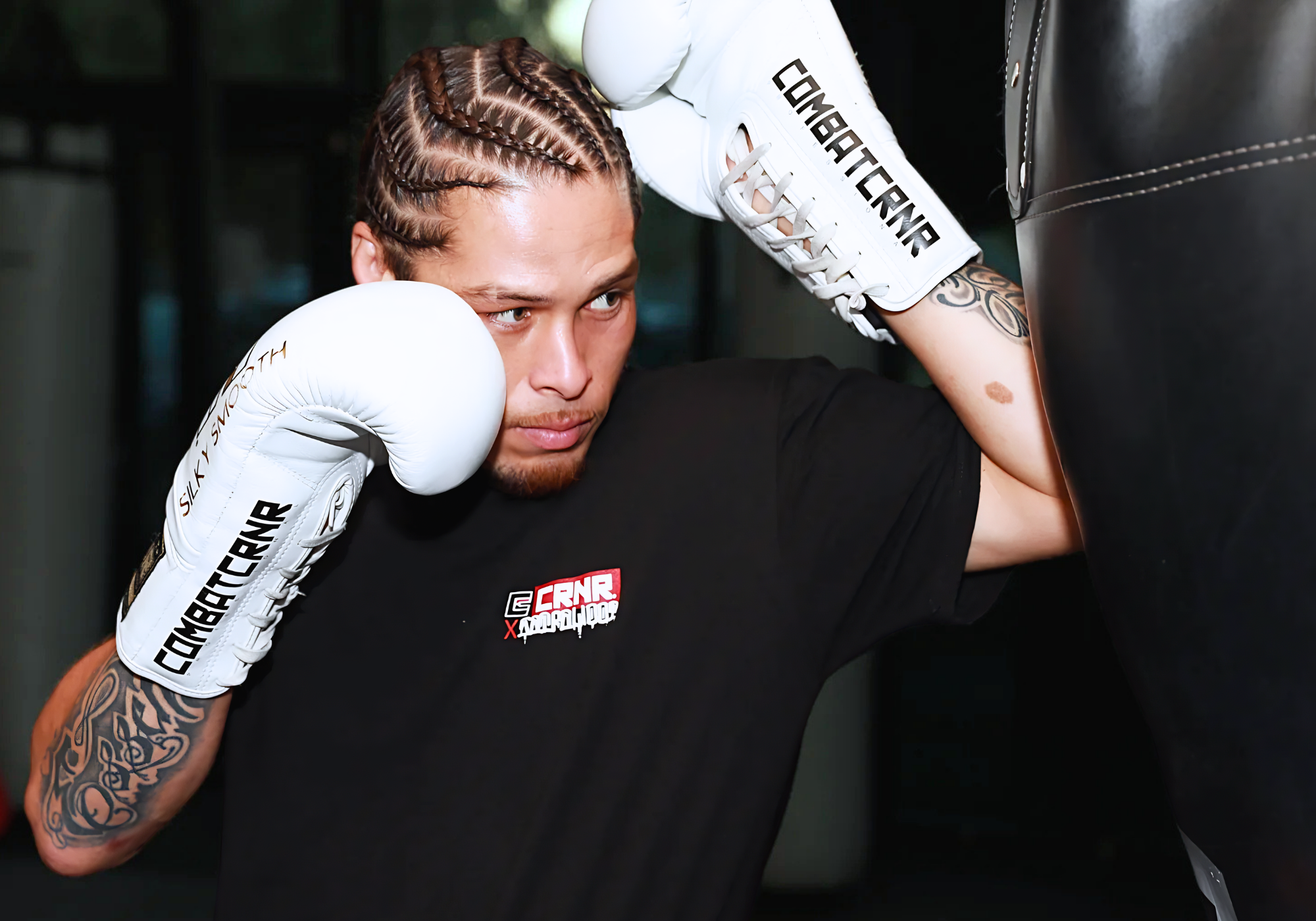Close-up shot of professional fighter Eddie Abasolo striking a heavy bag while wearing a black Combat Corner t-shirt and white lace-up Combat Corner boxing gloves with bold black lettering. 