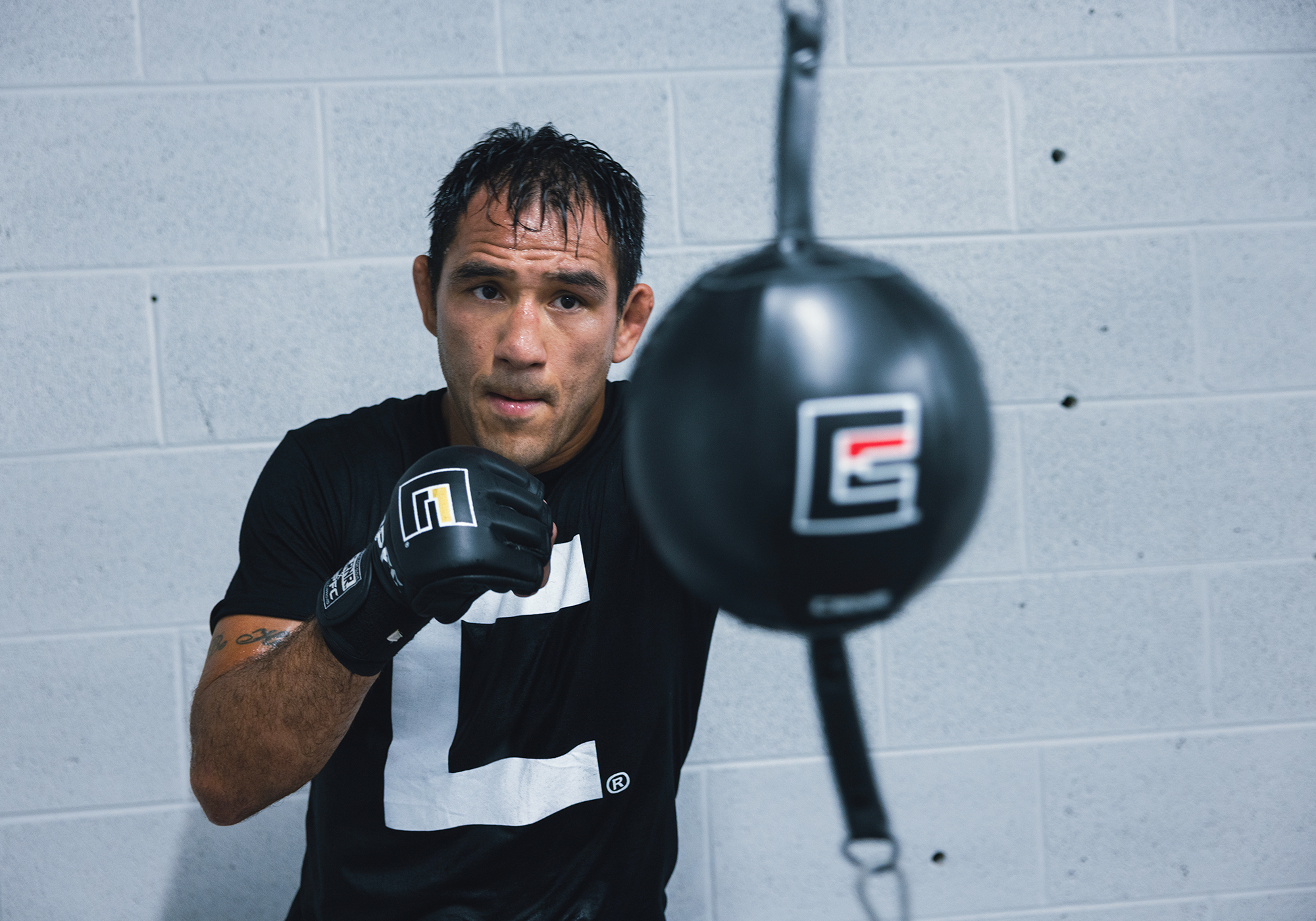 Professional fighter Emmanuel Sanchez training on a Combat Corner double-end bag in a gym, holding a boxing guard while wearing black Combat Corner MMA Gloves and a black Combat Corner Big C T-Shirt. 
