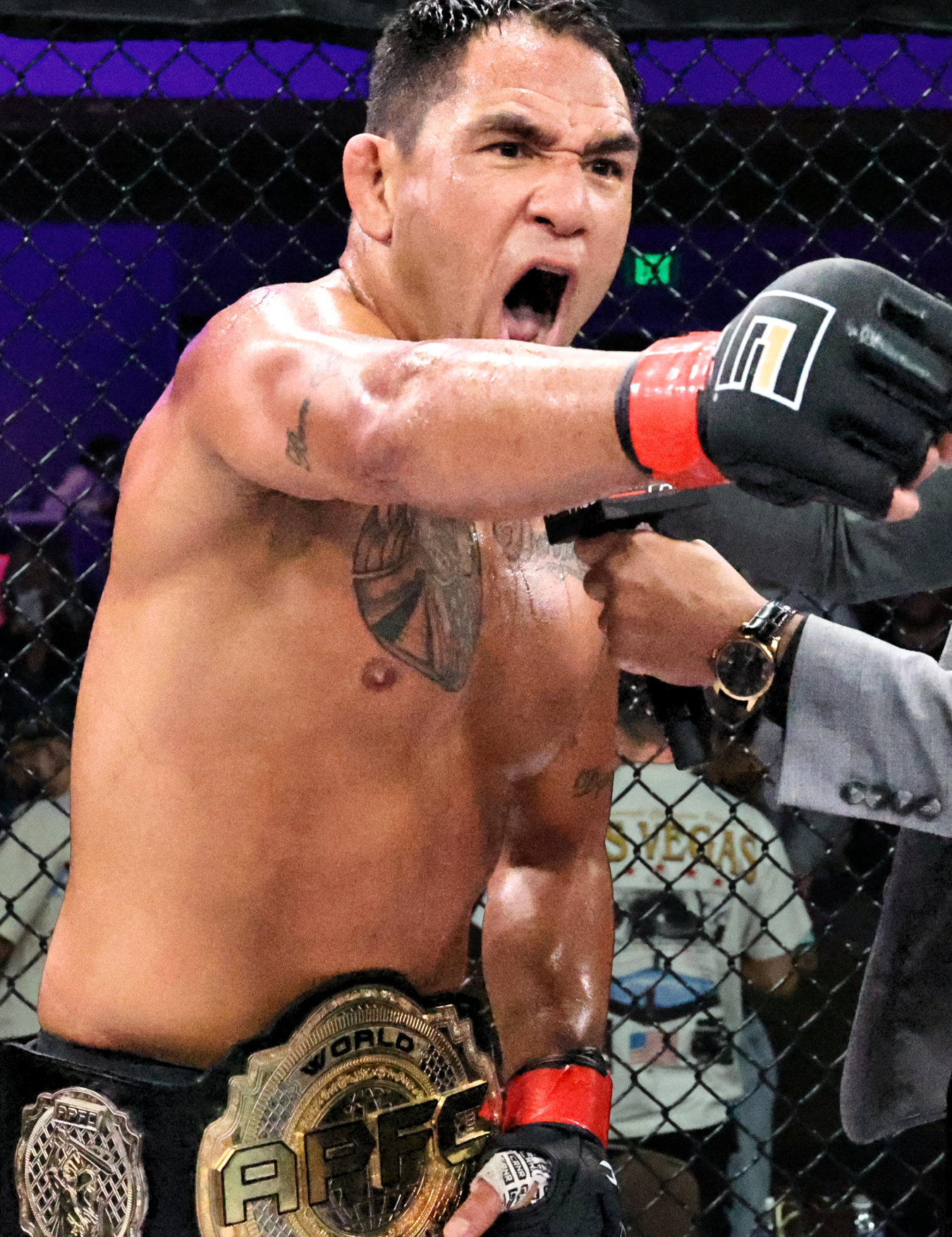 Close-up of a victorious APFC Champion and Combat Corner Pro Team Member Emmanuel Sanchez wearing a large gold APFC World Champion belt, punching directly toward the camera while wearing black Combat Corner APFC MMA Fight Gloves with a red wrist cuff. 