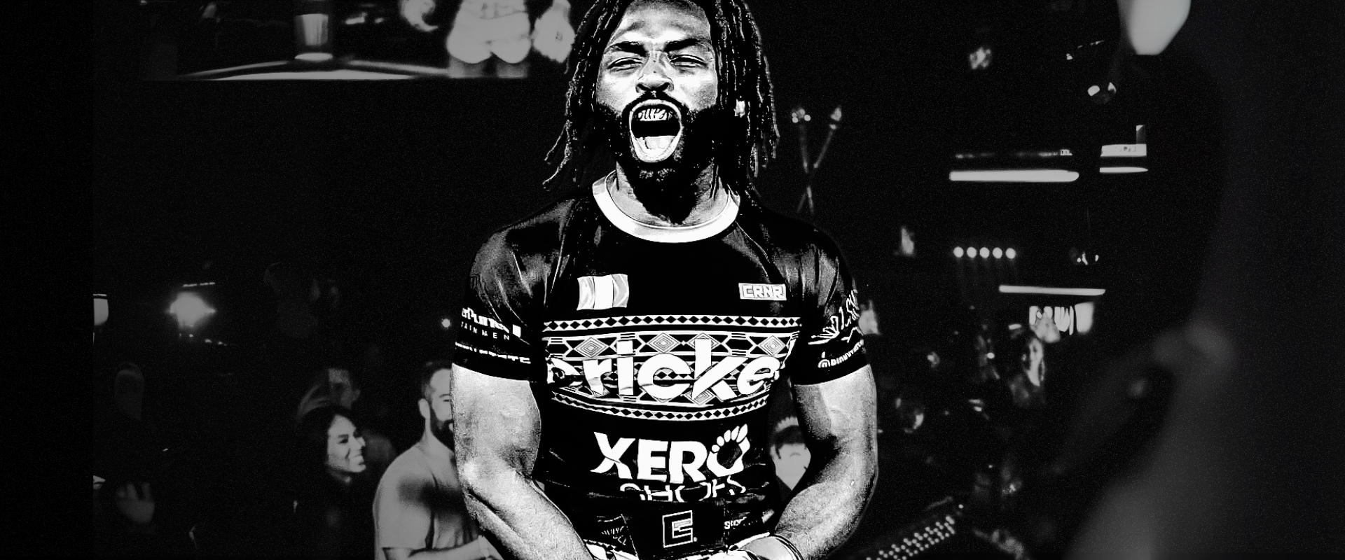 High-contrast black and white panoramic portrait of pro fighter and Combat Corner Pro Team Member Raufeon Stots with dreadlocks shouting in victory, wearing a custom graphic Combat Corner performance rash guard and fight shorts. 
